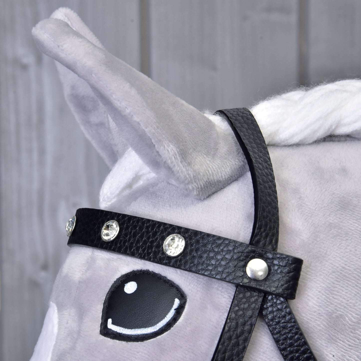 Detail hlavy šedého plyšového koně s Kompletní uzdečkou s kamínky z eko kůže od Hobby Horse LarDen. Kůň má bílou hřívu, vztyčené ucho, oko v kresleném stylu a v pozadí jsou světlé dřevěné desky.