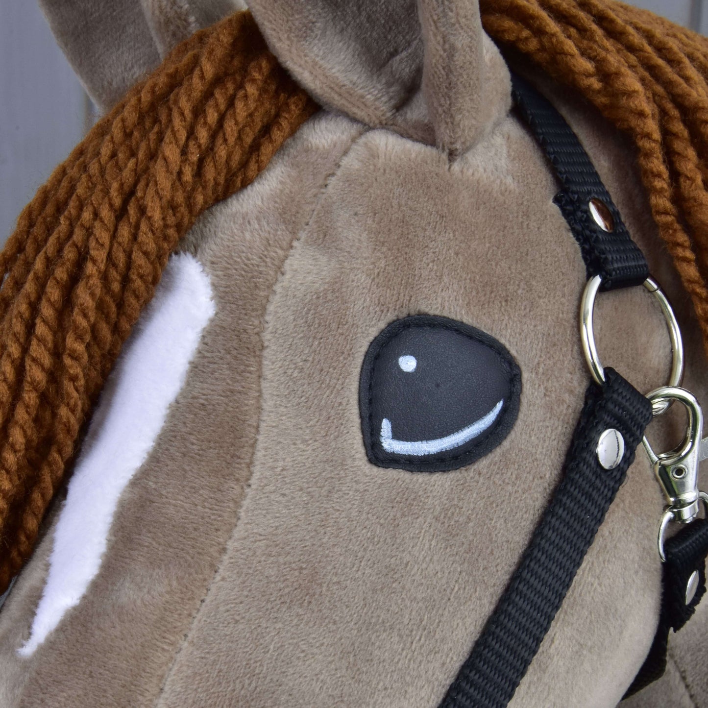 Detail Hobby Horse Oliver od Hobby Horse LarDen s čiernou ohlávkou a ohlávkou s barančekom zadarmo, s hnedou pletenou hrivou z priadze, bielou škvrnou okolo oka a našitým okom v kreslenom štýle, so vzhľadom roztomilého drezúrneho koňa.