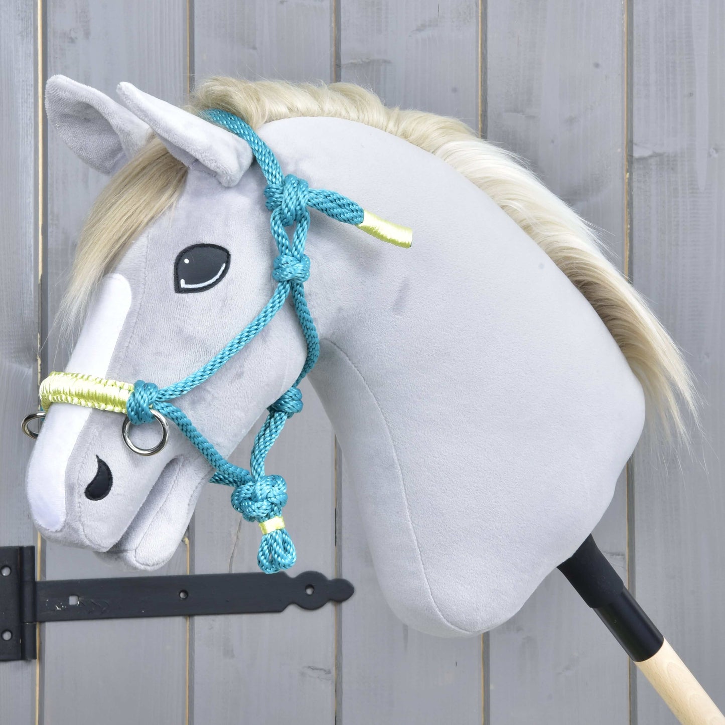 Hobby Horse LarDen Parelka Tyrkys na plyšovom Hobby Horse so svetlosivou hlavou, bielou hrivou a modrou uzdečkou, nasadenom na drevenej tyči pred sivou drevenou stenou.