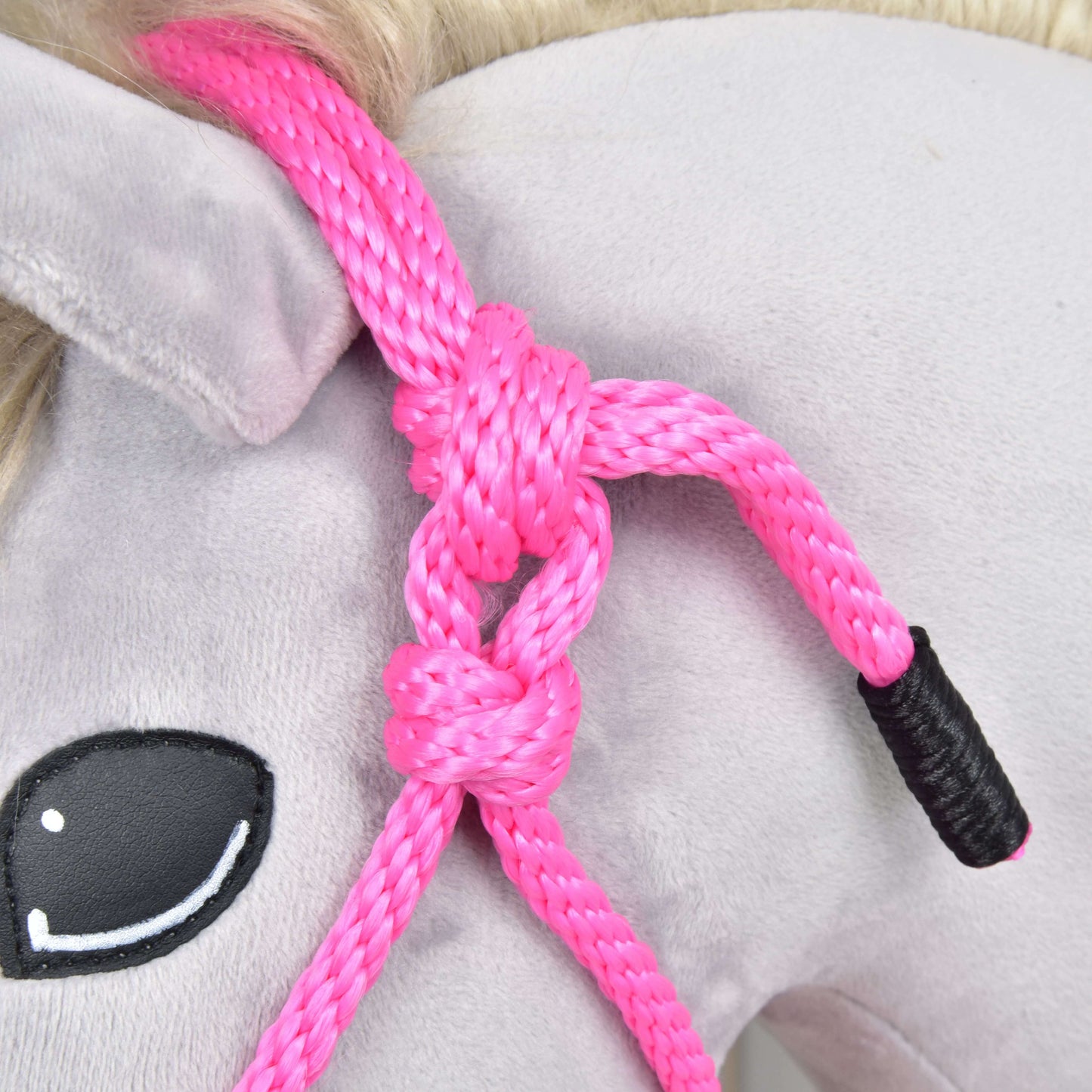 Detail hlavy plyšového koňa s Hobby Horse LarDen Parelka Pink, uviazanou okolo ucha. Kôň má svetlosivé telo, maľované čierne oko s bielymi odleskami a svetlú blond hrivu.