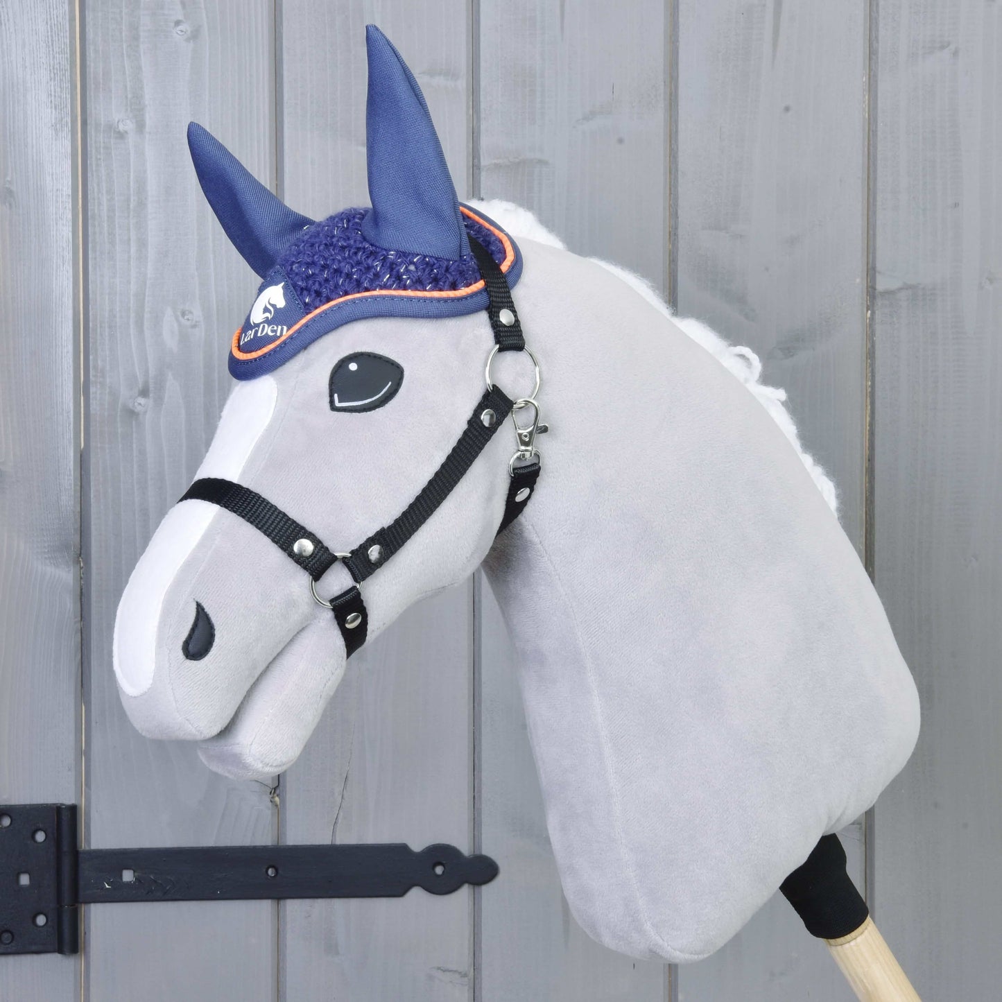 Čabraka háčkovaná Blue sport od Hobby Horse LarDen je zobrazena na plyšovém Hobby Horse s bílým obličejem a černou uzdečkou, nasazeném na dřevěné tyči před šedými panelovými dveřmi s černým pantem.