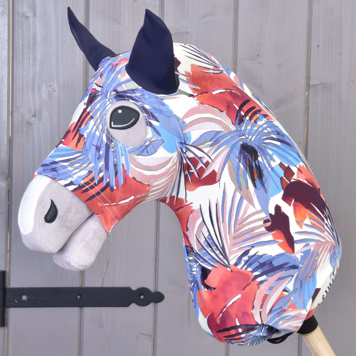 Hobby Horse ochranný oblek Blue Jungle (Lycra) od Hobby Horse LarDen má výrazný abstraktný vzor v červenej, bielej, fialovej a modrej farbe s čiernymi očami a tmavými ušami. Obsahuje prateľný poťah a je zobrazený na drevenej tyči pri sivej stene.