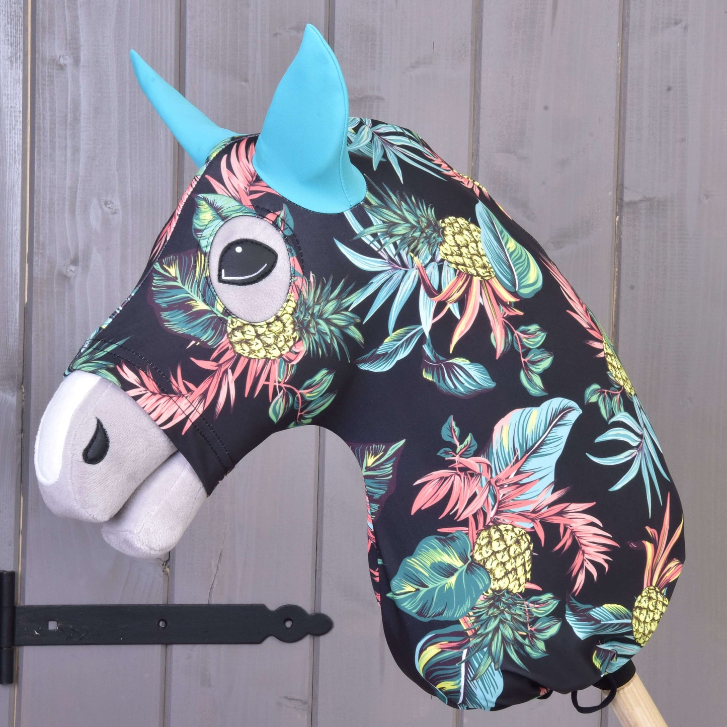 Hobby Horse ochranný oblek Pineapple (Lycra) od Hobby Horse LarDen má čiernu hlavu s výraznou potlačou ananásov a listov, modré uši a biely ňufák, je nasadený na drevenej tyči a zobrazený pred sivou drevenou stenou.