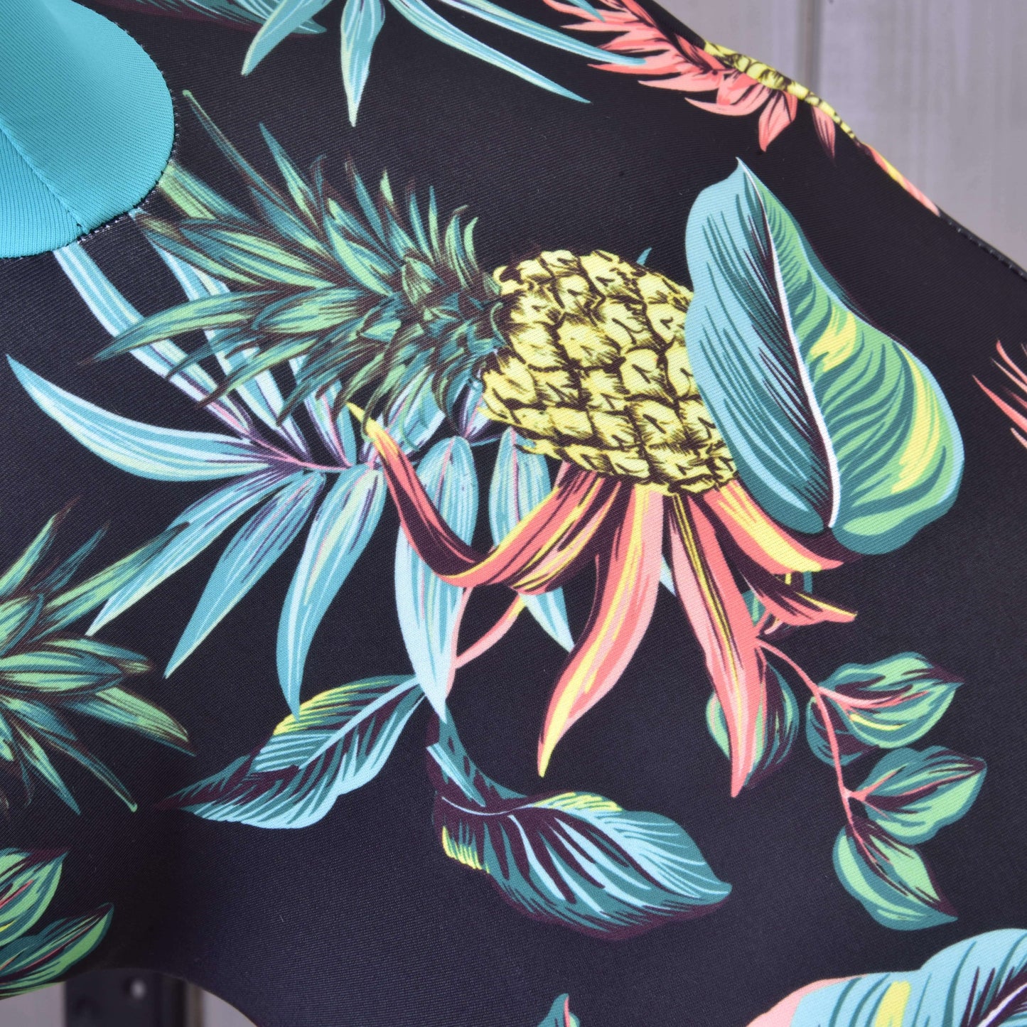 Detail Hobby Horse ochranný oblek Pineapple (Lycra) od Hobby Horse LarDen, vyrobeného z PES a Lycra, s výraznými ananásmi, zelenými listami a tropickým porastom na čiernom podklade, ideálny pre vytvorenie štýlového ochranného obleku.