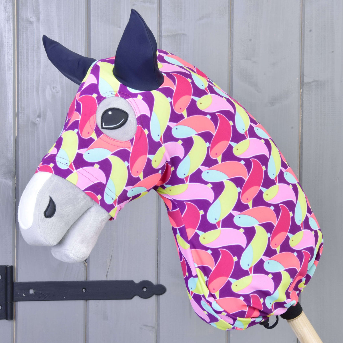 Hobby Horse ochranný oblek Bird (Lycra) od Hobby Horse LarDen má výrazný lycrový poťah so vzorom vtákov v ružovej, žltej, zelenej a modrej farbe na drevenej tyči a je zobrazený na pozadí sivej drevenej steny.