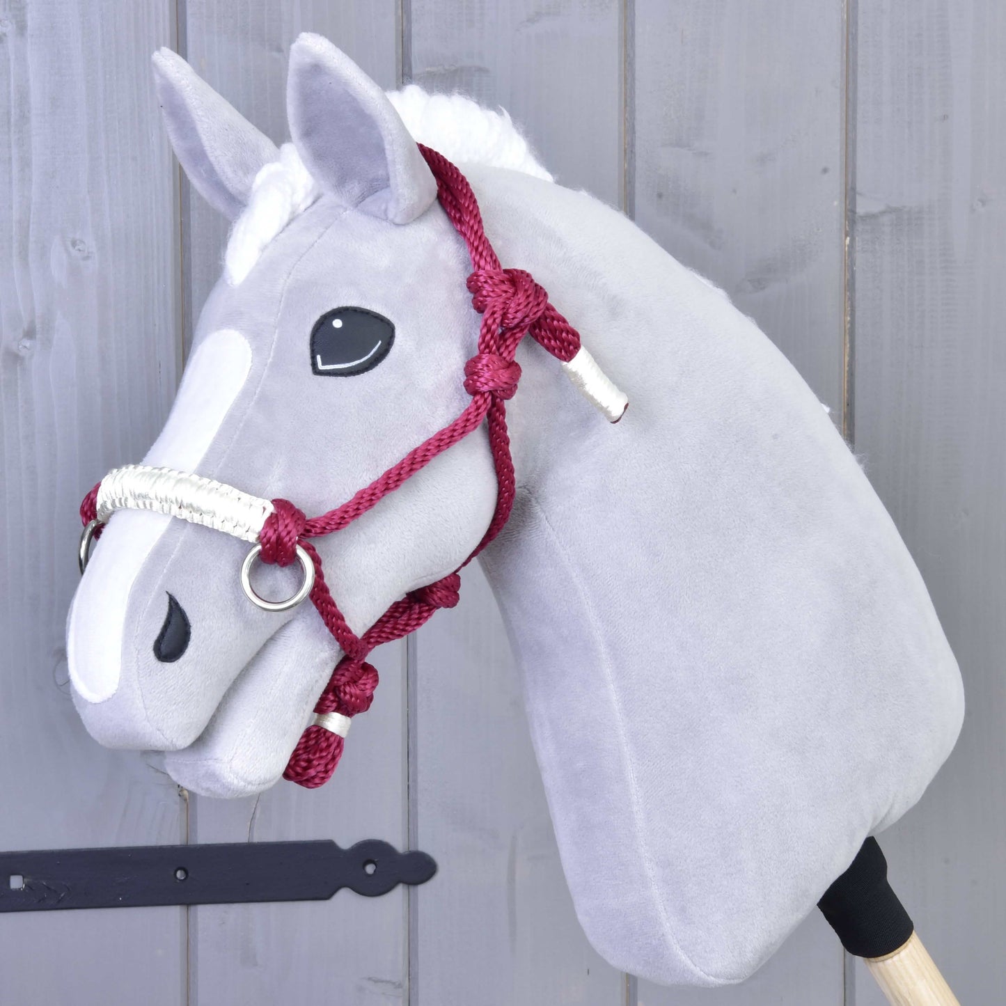 Sivý plyšový Hobby Horse od Hobby Horse LarDen s bielou lysinou, čiernymi gombíkovými očami a doplnkom Parelka Czi-Czi, na drevenej tyči pred sivou drevenou stenou.