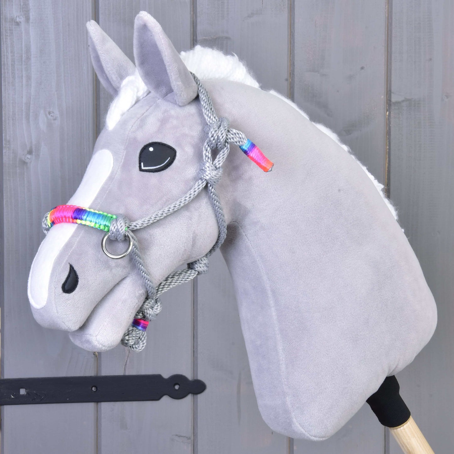 Hobby Horse LarDen Parelka Manhattan na mäkkom sivom doplnku pre Hobby Horse s bielou lysinou, čiernymi gombíkovými očami a pletenou povrazovou ohlávkou s dúhovými akcentmi, na sivom drevenom pozadí.