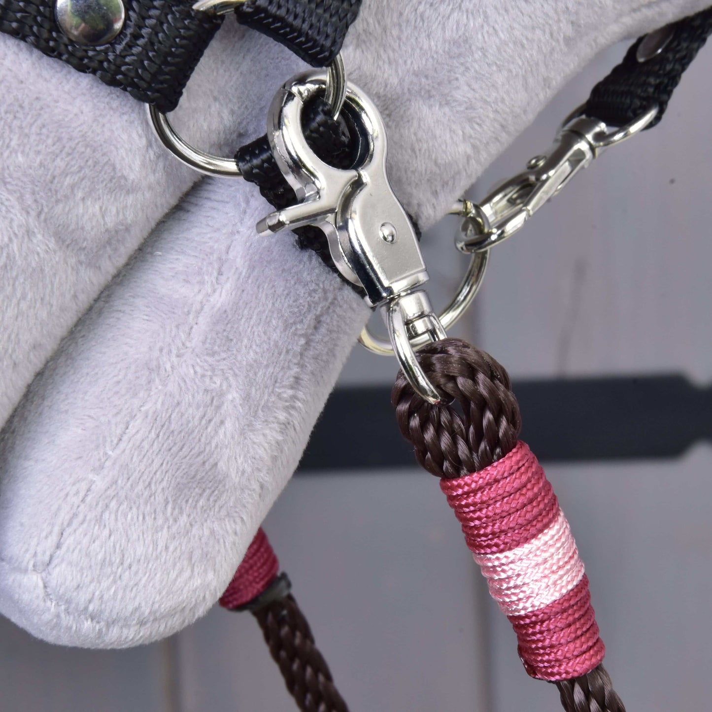 Detail kovového klipu pripevňujúceho Hobby Horse LarDen Opraty York — hnedé a ružové pletené polypropylénové lano — k čiernemu textilnému popruhu na mäkkom svetlosivom povrchu, pravdepodobne ako súčasť výbavy pre Hobby Horse.