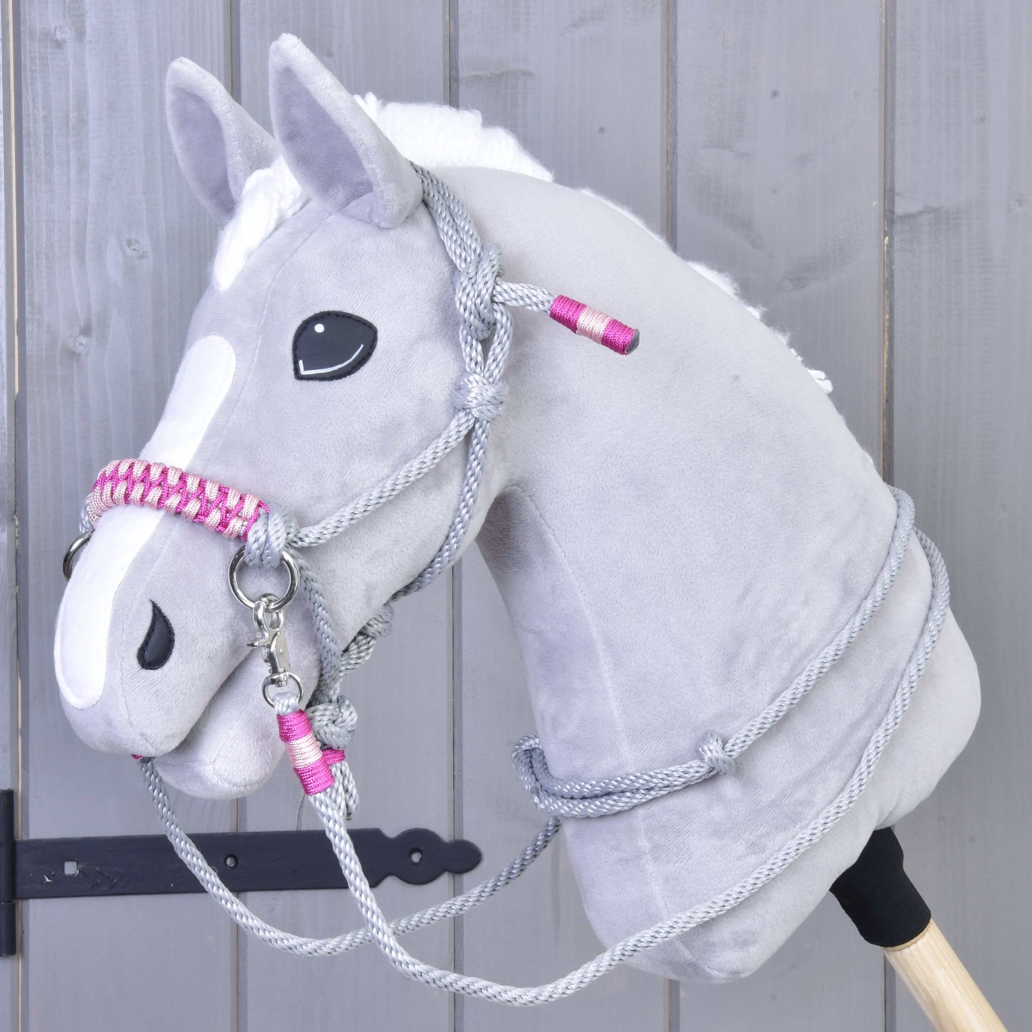 Povrazový set Rosie od Hobby Horse LarDen, sivý plyšový Hobby Horse s bielou lysinou, čiernymi očami a ružovo-sivou uzdečkou s opratami na drevenej tyči, je zobrazený pred svetlosivou drevenou stenou.