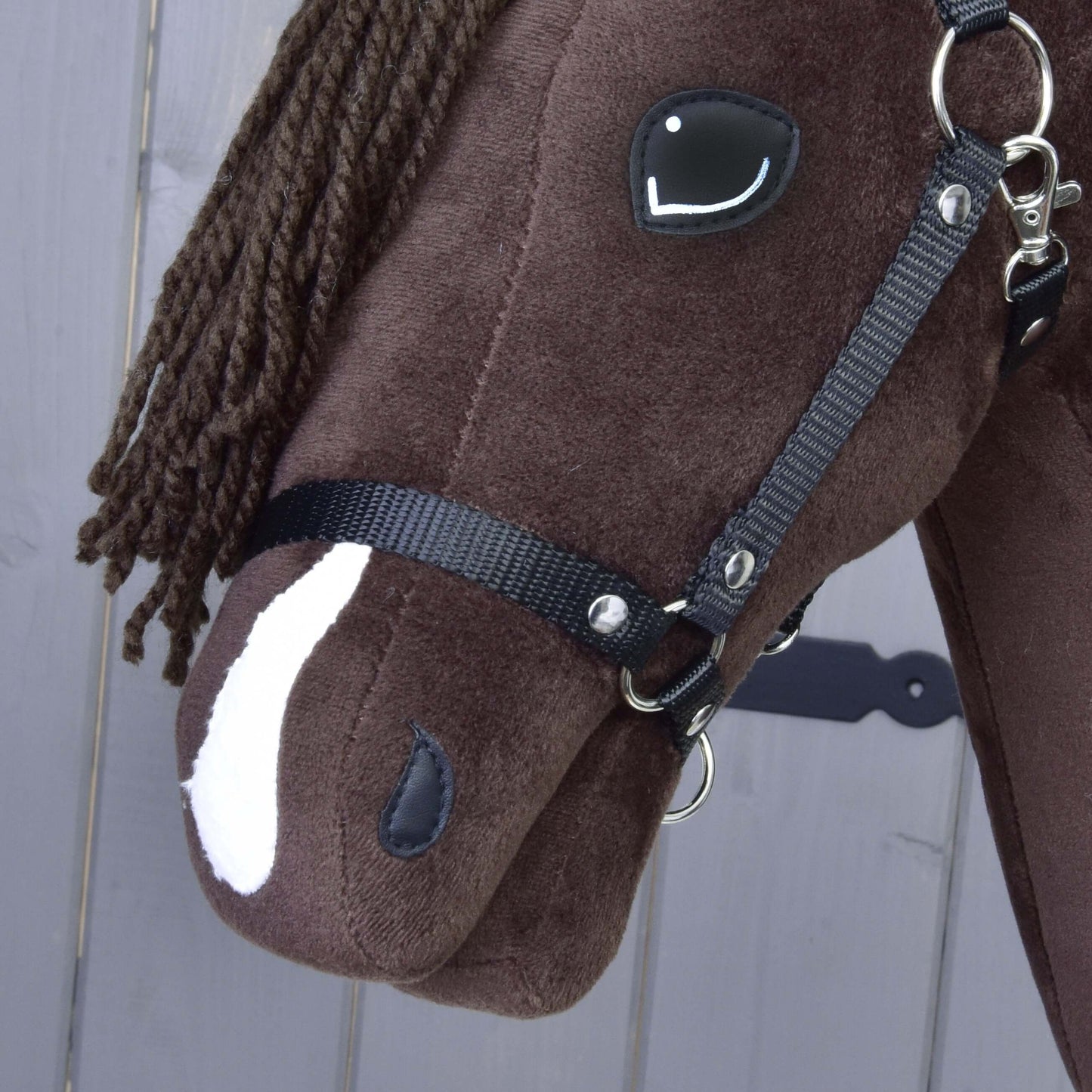 Detail Hobby Horse Chianti s čiernou ohlávkou od Hobby Horse LarDen, inšpirovaného American Quarter Horse, s čiernou priadzovou hrivou, bielou škvrnou na nose a čiernou uzdečkou pred sivým dreveným pozadím.