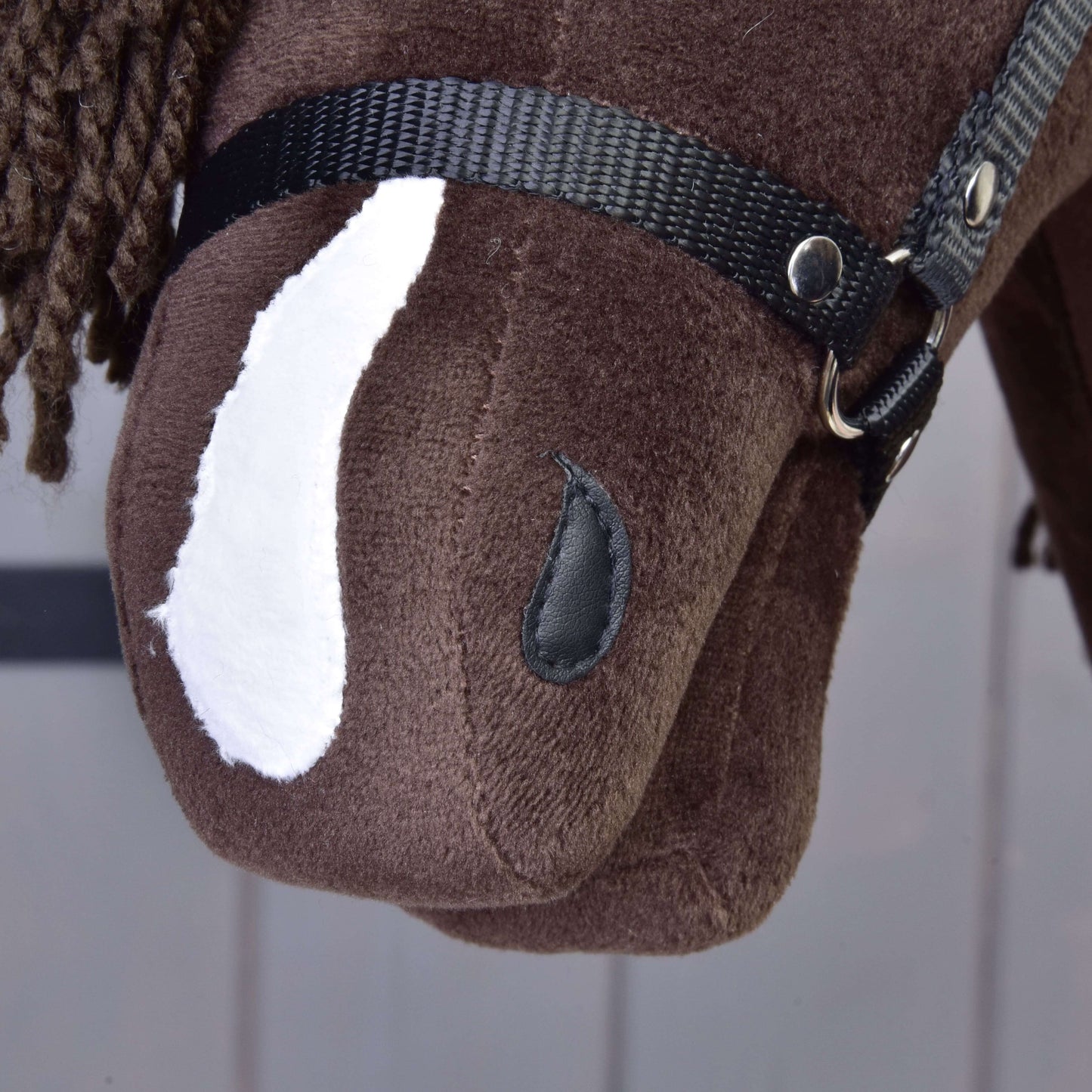 Detail Hobby Horse Chianti s čiernou ohlávkou od Hobby Horse LarDen: hnedá látková hlava koňa s bielou škvrnou, čiernou ohlávkou, gombíkovým okom a hnedou priadzovou hrivou na jemne pruhovanom svetlom pozadí.