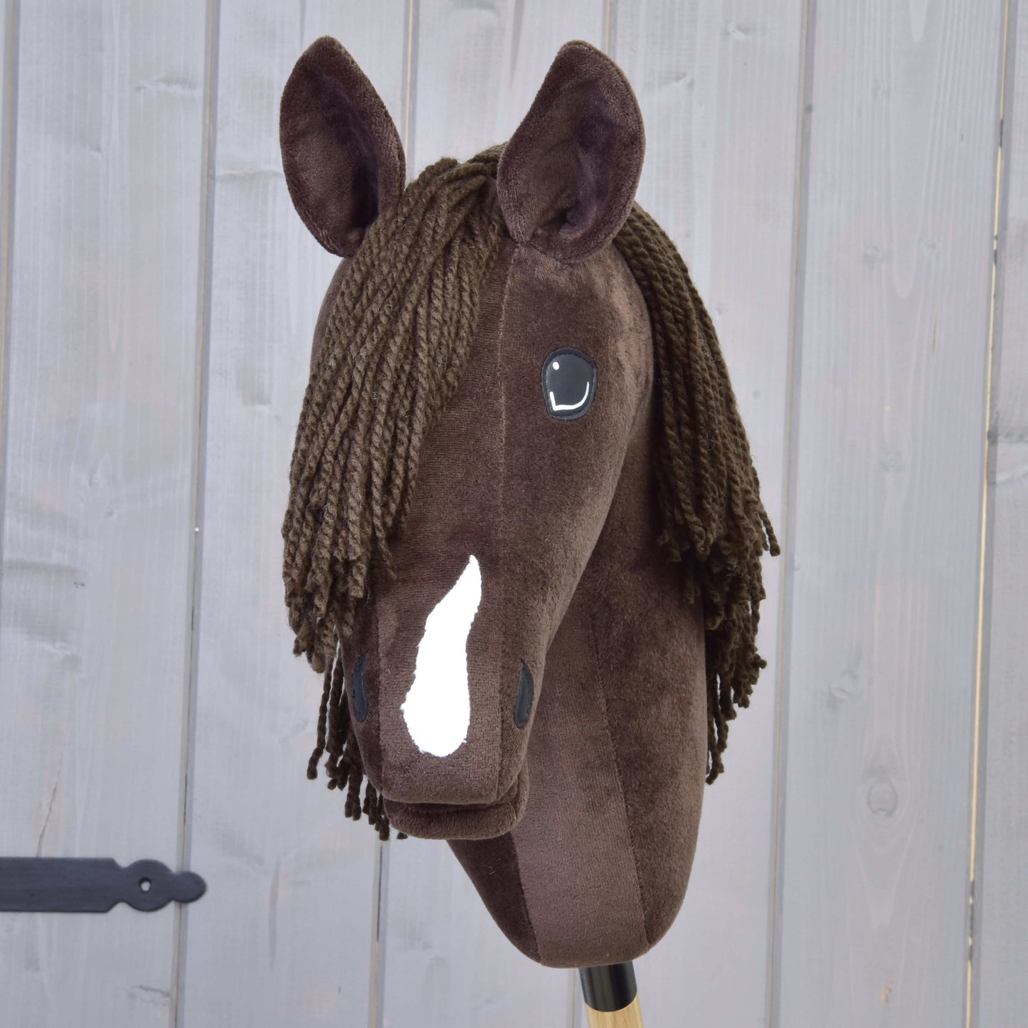Hobby Horse Chianti s čiernou ohlávkou od Hobby Horse LarDen je hnedý plyšový Hobby Horse s bielou lysinou, čiernymi gombíkovými očami a hnedou priadzovou hrivou, inšpirovaný pokojným American Quarter Horse. Na pozadí drevených panelov.