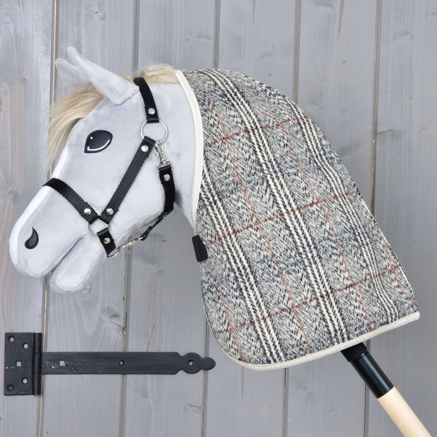 Sivý plyšový Hobby Horse od Hobby Horse LarDen s čiernou uzdečkou a dekou Deka stajňová Fine Check, nasadený na drevenej tyči pred svetlosivou drevenou stenou.