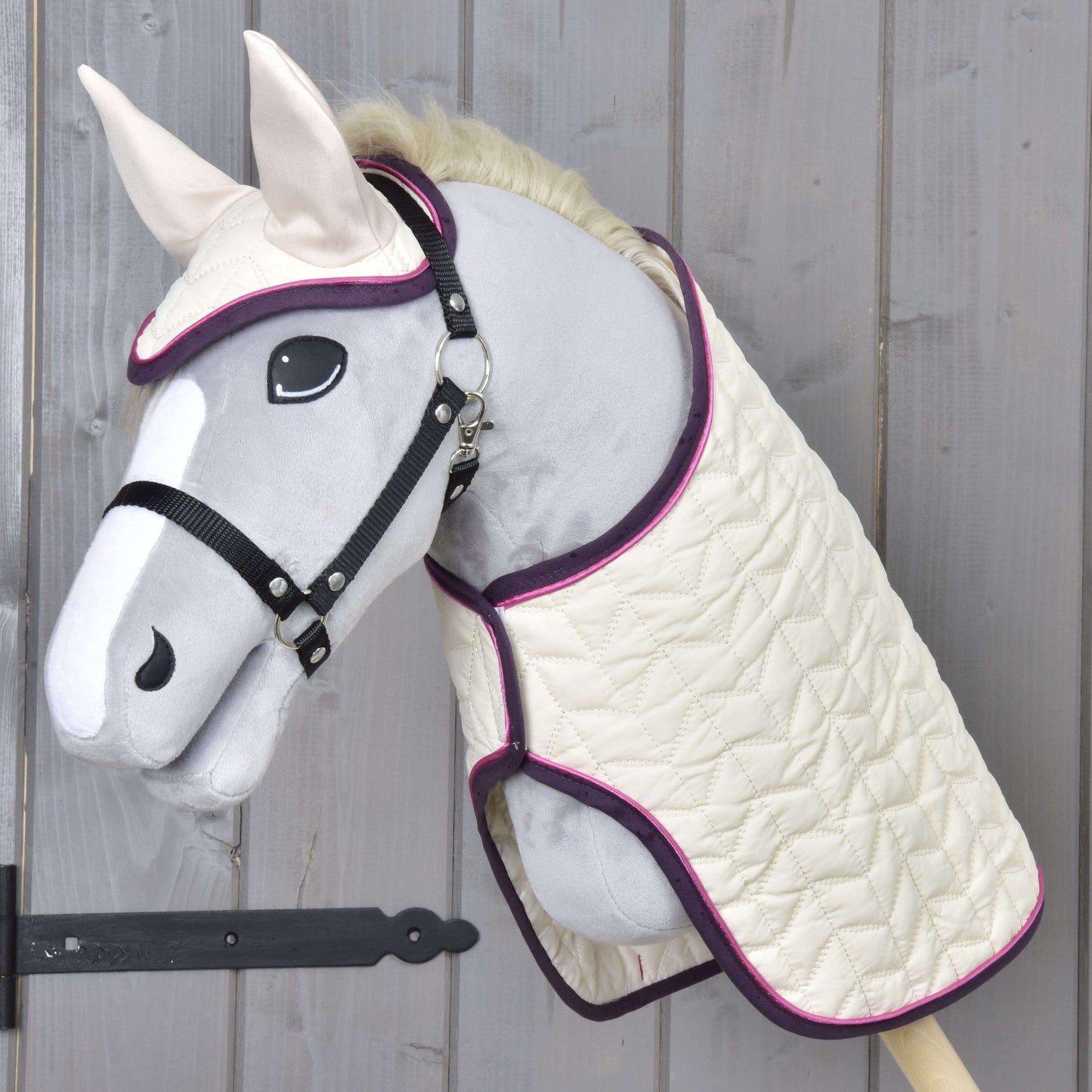 Set Supreme Crema od Hobby Horse LarDen s plyšovým Hobby Horse se šedým obličejem, bílou tlamou a ušima, oblečeným do prošívané krémové deky a ladící čabraky s fialovým lemem, vystavený na dřevěné stěně.