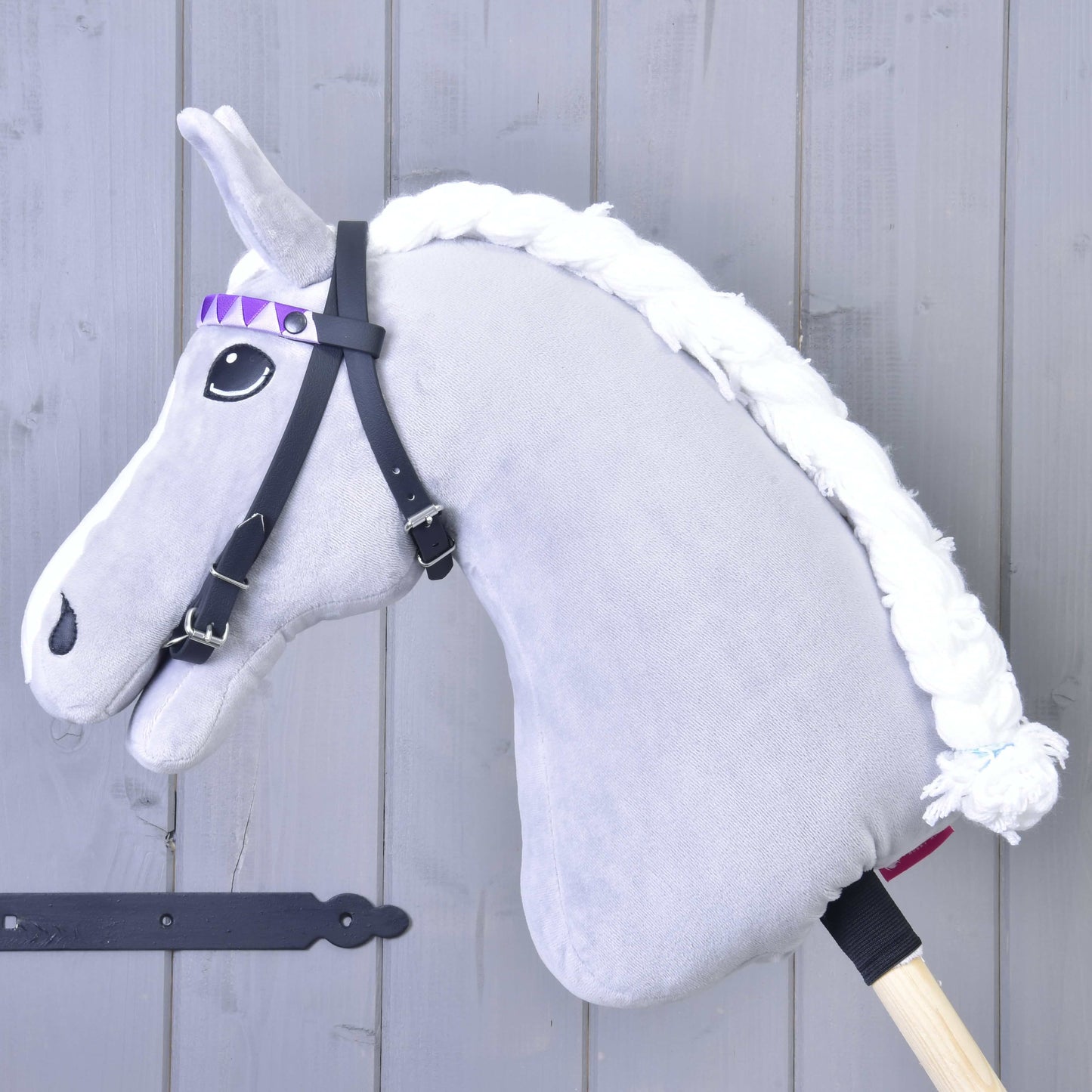 Uzdečka bez nánosníku fialová se 2 čelenkami (vel. M) od Hobby Horse LarDen, se šedým plyšovým koněm s bílou pletenou hřívou, černou uzdečkou bez nánosníku a fialovou stuhou s kosočtvercovým vzorem na dřevěné tyči, u světle šedé dřevěné stěny.