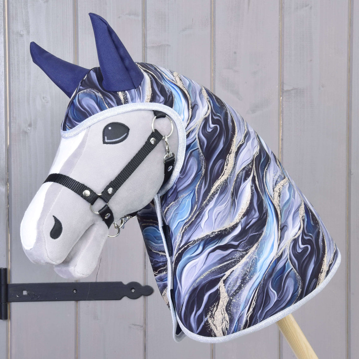 Pláštenka Galaxy Cobalt od Hobby Horse LarDen s mramorovanou modrou, bielou a čiernou látkou, tmavomodrými ušami, čiernou ohlávkou a ladiacimi doplnkami na drevenej tyči, na pozadí svetlej drevenej steny.