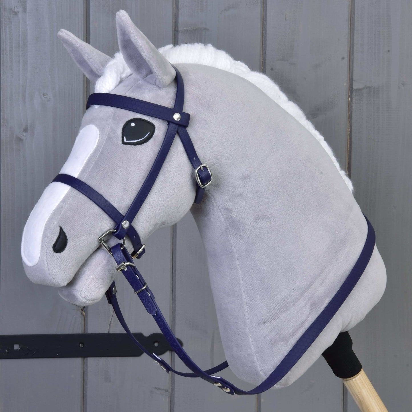 Plyšový sivý Hobby Horse od Hobby Horse LarDen s kompletnou uzdečkou s nánosníkom v tmavofialovej farbe, čierno-bielymi očami, bielou hrivou, nasadený na drevenej tyči, stojí pred sivou drevenou stenou.