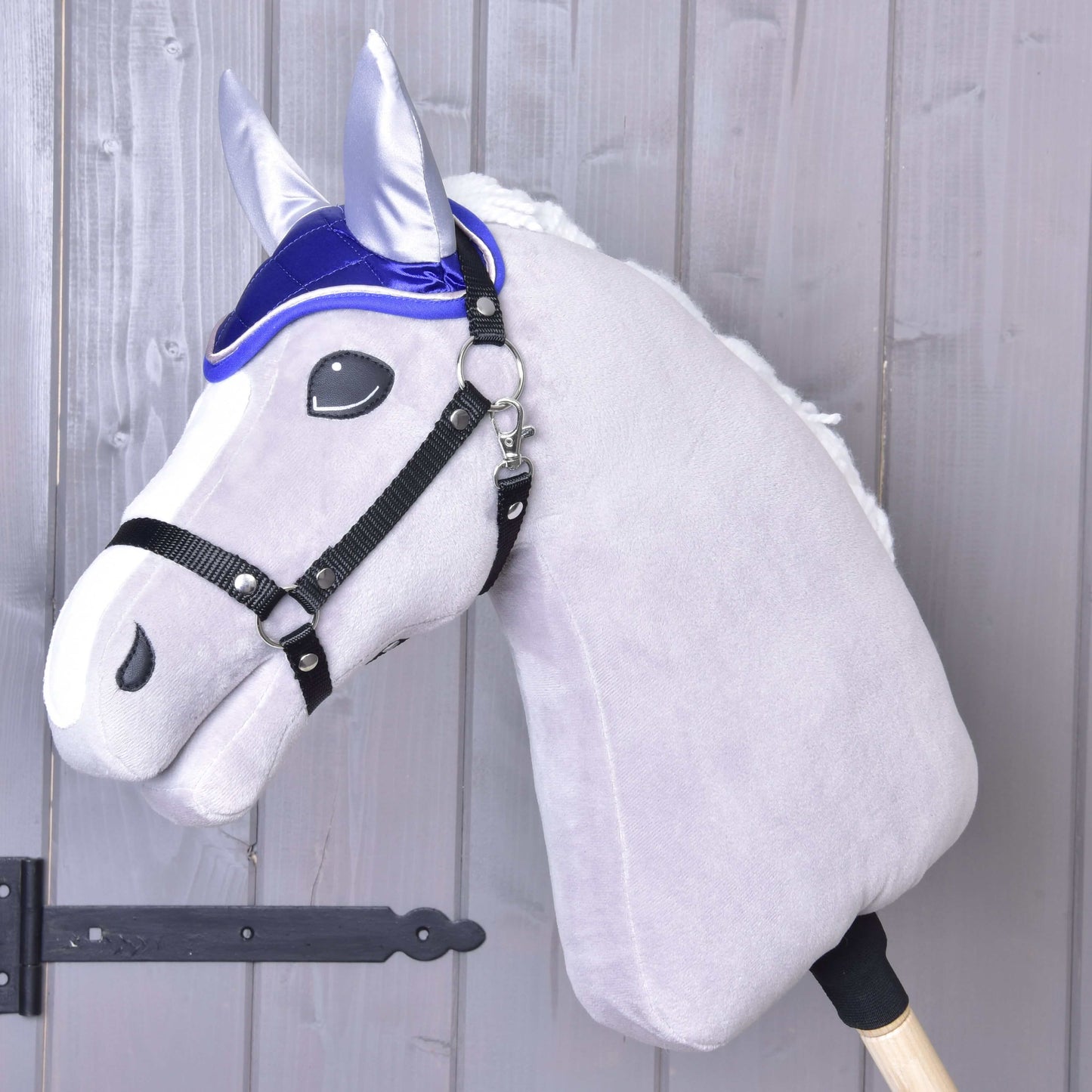 Plyšový Hobby Horse od Hobby Horse LarDen s bielou hrivou, čiernou uzdečkou a čabrakou DeLuxe Blue pre ochranu proti hmyzu, nasadený na drevenej tyči pred svetlosivou drevenou stenou.