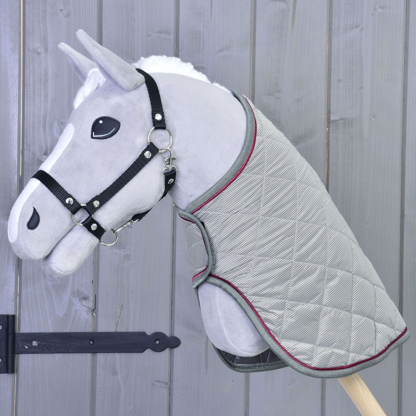 Sivý plyšový Hobby Horse od Hobby Horse LarDen s čiernou uzdečkou a dekou Silver Eleganza stojí pred drevenou stenou so zvislými panelmi.