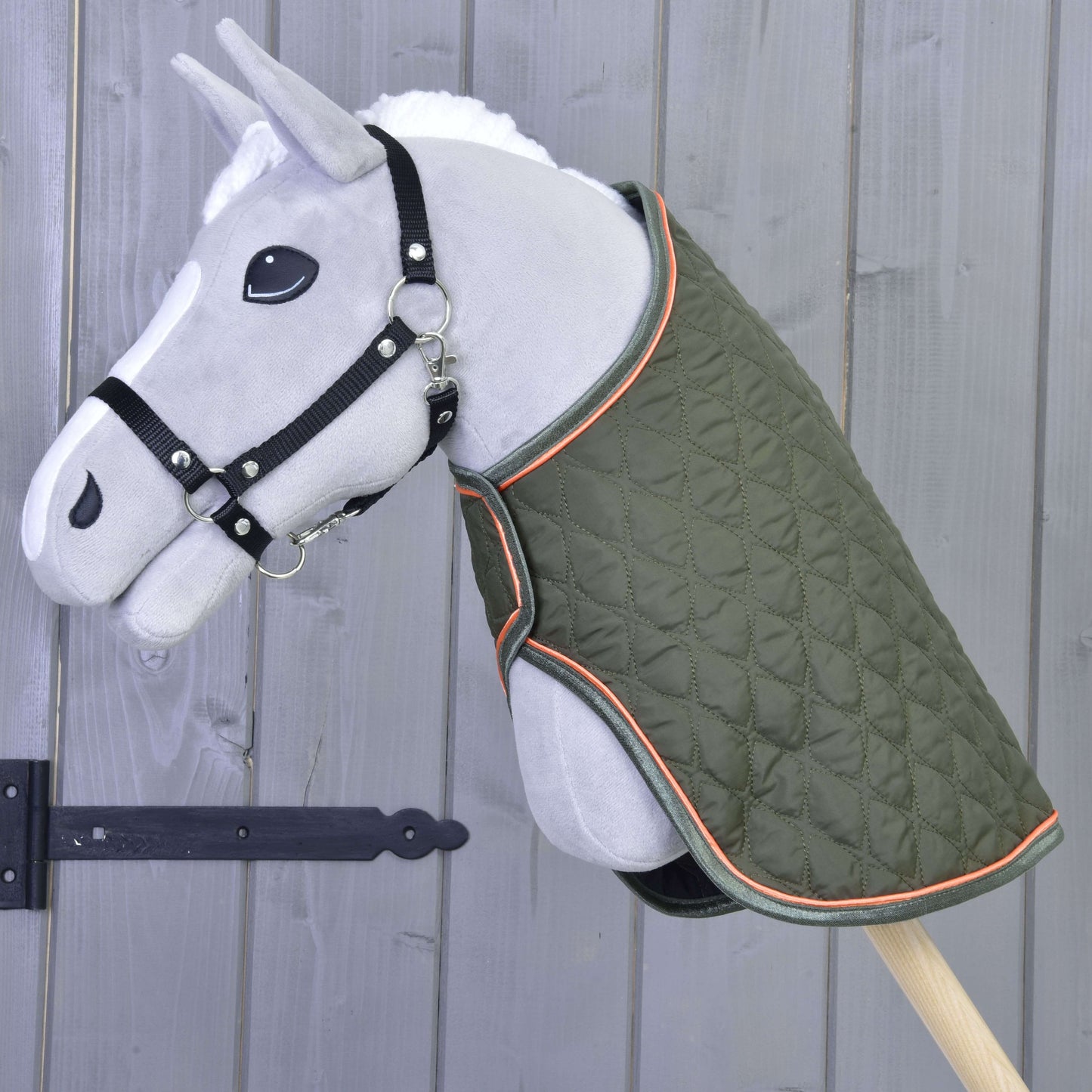 Sivý plyšový Hobby Horse na tyči s Dekou Sport od Hobby Horse LarDen v zelenej farbe s oranžovým lemovaním a čiernou ohlávkou stojí pred drevenou stenou s čiernym železným závesom.