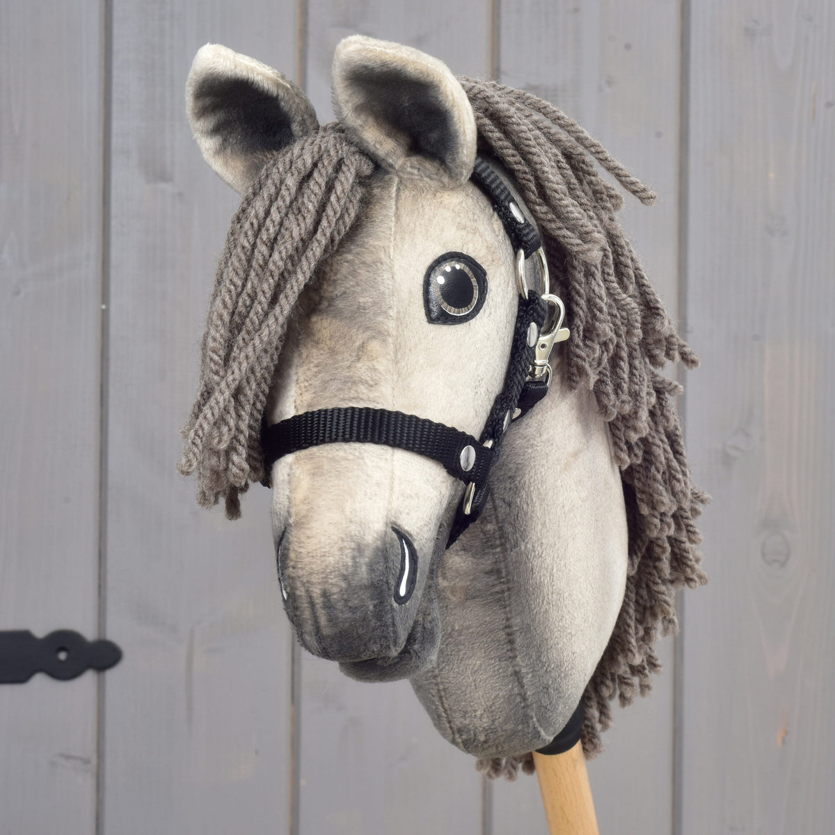 Detailný záber na Hobby Horse Dusty s čiernou ohlávkou vo veľkosti XS od Hobby Horse LarDen podľa hannoverského žrebca, so sivou priadzovou hrivou, čiernou ohlávkou a výraznými gombíkovými očami, nasadený na drevenej tyči na pozadí sivého dreva.