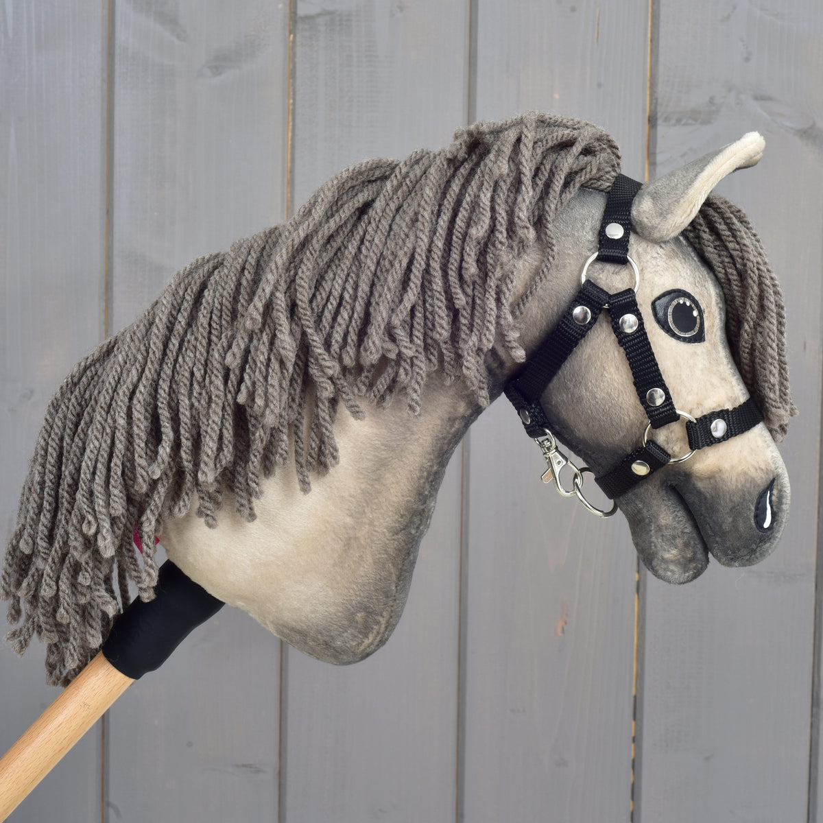 Hobby Horse Dusty s čiernou ohlávkou vo veľkosti XS od Hobby Horse LarDen so sivou priadzovou hrivou a drevenou tyčou ako držadlom je zobrazený pred sivým dreveným plotom.