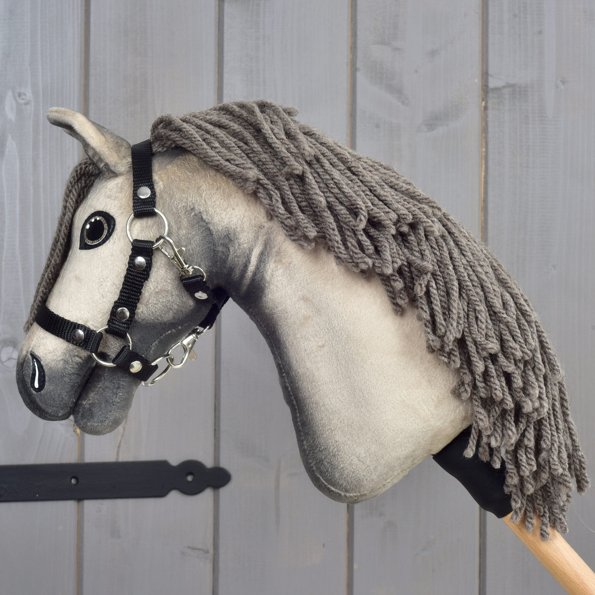 Hobby Horse Dusty s čiernou ohlávkou vo veľkosti XS od Hobby Horse LarDen so sivou srsťou, priadzovou hrivou a čiernou ohlávkou je nasadený na drevenej tyči a umiestnený pred svetlosivou drevenou stenou.