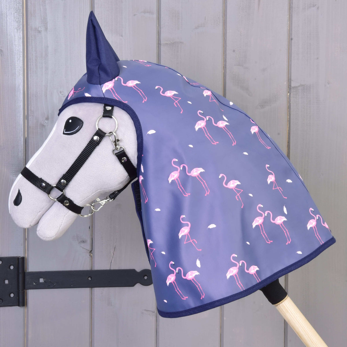 Sivý Hobby Horse LarDen s čiernou uzdečkou má na sebe Pláštenku Flamingo – modrú kapucňu do každého počasia s ružovými plameniakmi a bielymi bodkami – na drevenom pozadí.