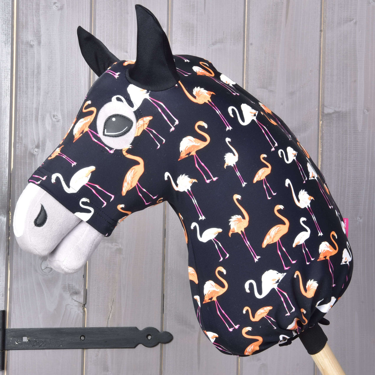 Hobby Horse Lycra ochranný oblek Flamingo od Hobby Horse LarDen s lycrovým poťahom s oranžovými a bielymi plameniakmi na tmavom podklade, nasadený na drevenej tyči pred stenou z drevených panelov.