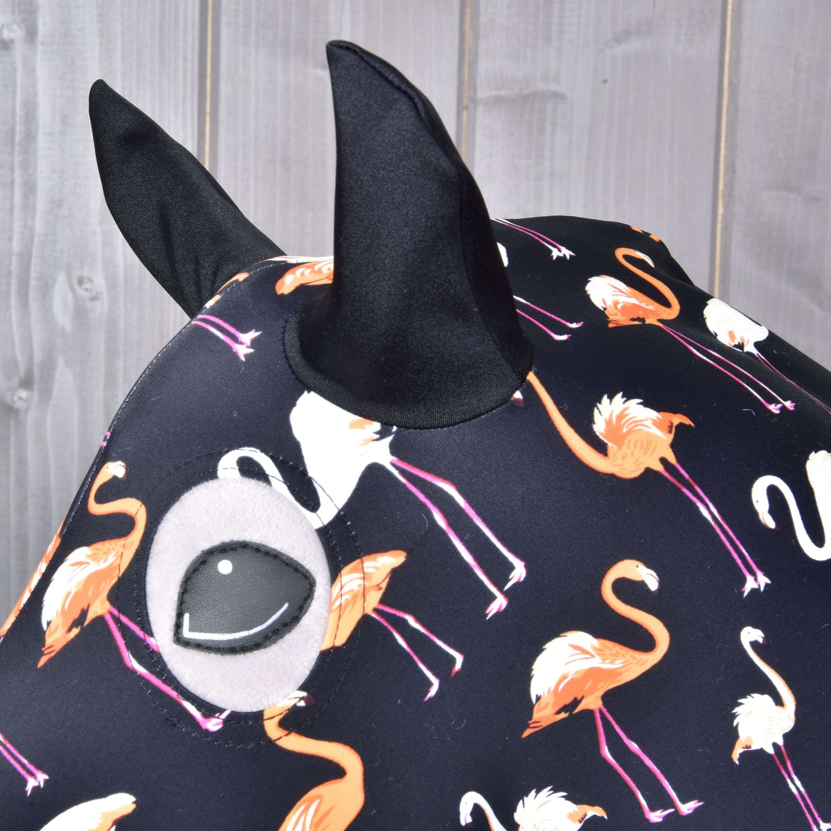 Detail Hobby Horse Lycra ochranného obleku Flamingo od Hobby Horse LarDen s oranžovými a bielymi plameniakmi na tmavom podklade, čiernymi špicatými ušami a kresleným okom. Za látkovým objektom vidno drevené panely.