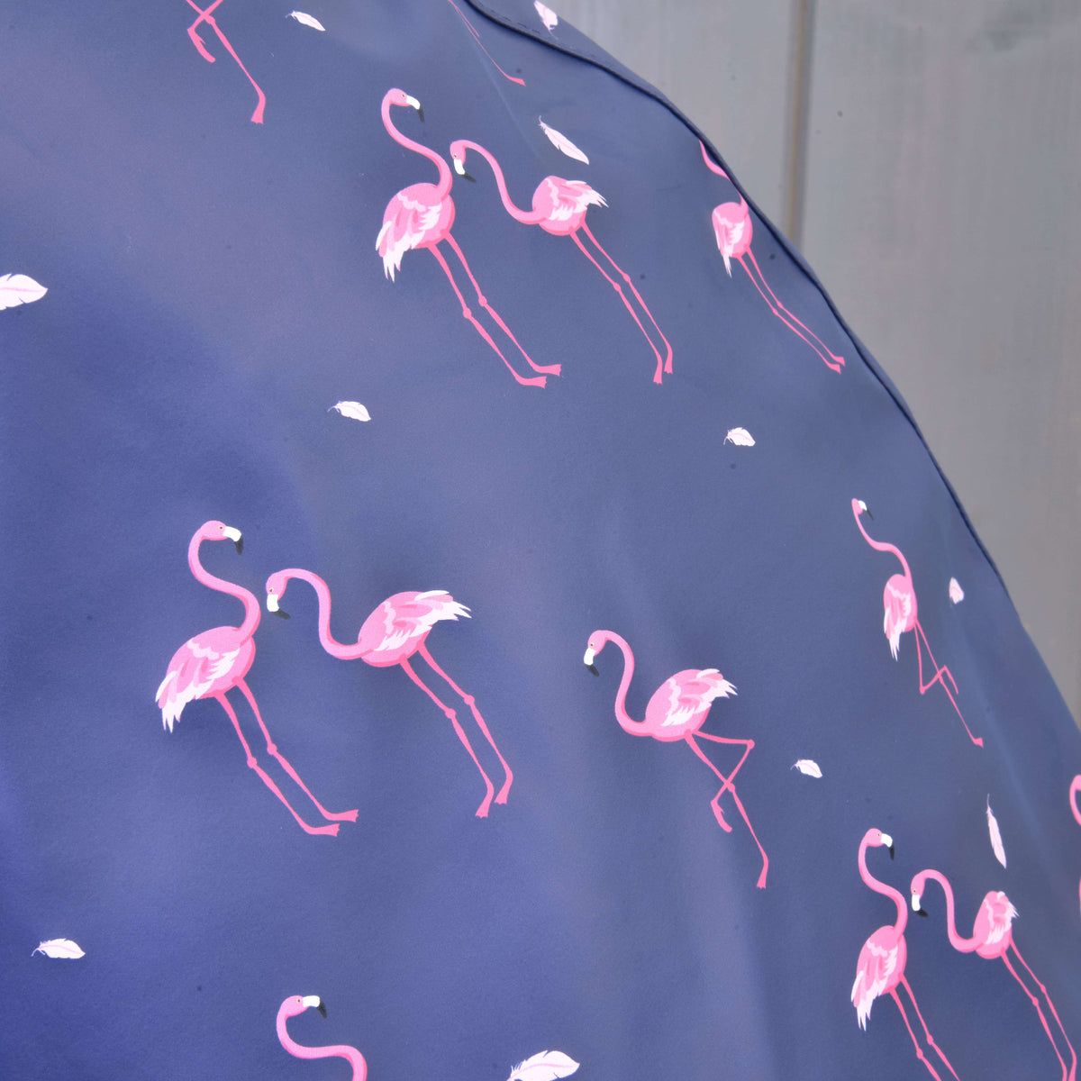 Detail Pláštenky Flamingo od Hobby Horse LarDen ukazuje modrú látku s ružovými plameniakmi a bielymi perami na jemne rozostrenom pozadí – ideálna do každého počasia alebo pre hravé dni.