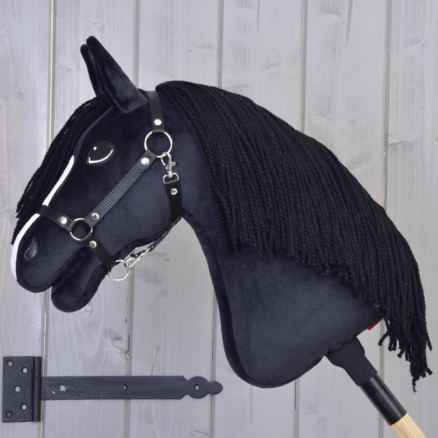 Hobby Horse Freya s černou ohlávkou od Hobby Horse LarDen s bílým pruhem na obličeji a černou přízovou hřívou na dřevěné tyči je umístěná před světle šedou dřevěnou stěnou – ideální pro každého rodinného nadšence do koní.