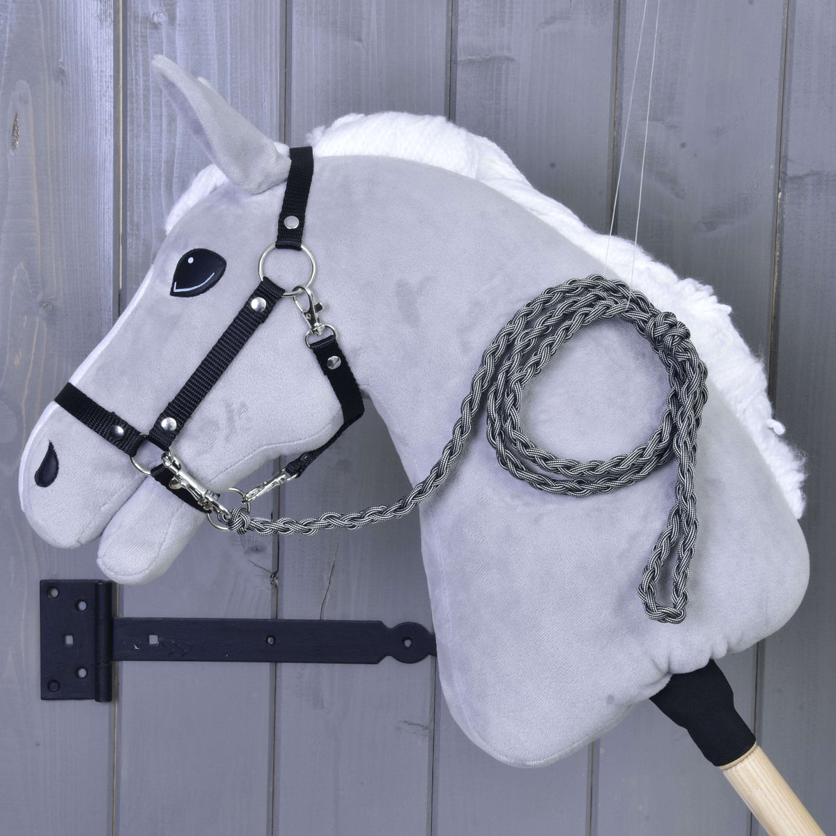 Hobby Horse - pletené vodidlo Graphite od Hobby Horse LarDen so sivou plyšovou hlavou, čiernou uzdečkou a ručne pleteným grafitovým vodidlom na drevenej tyči, zobrazené pred sivou drevenou stenou.