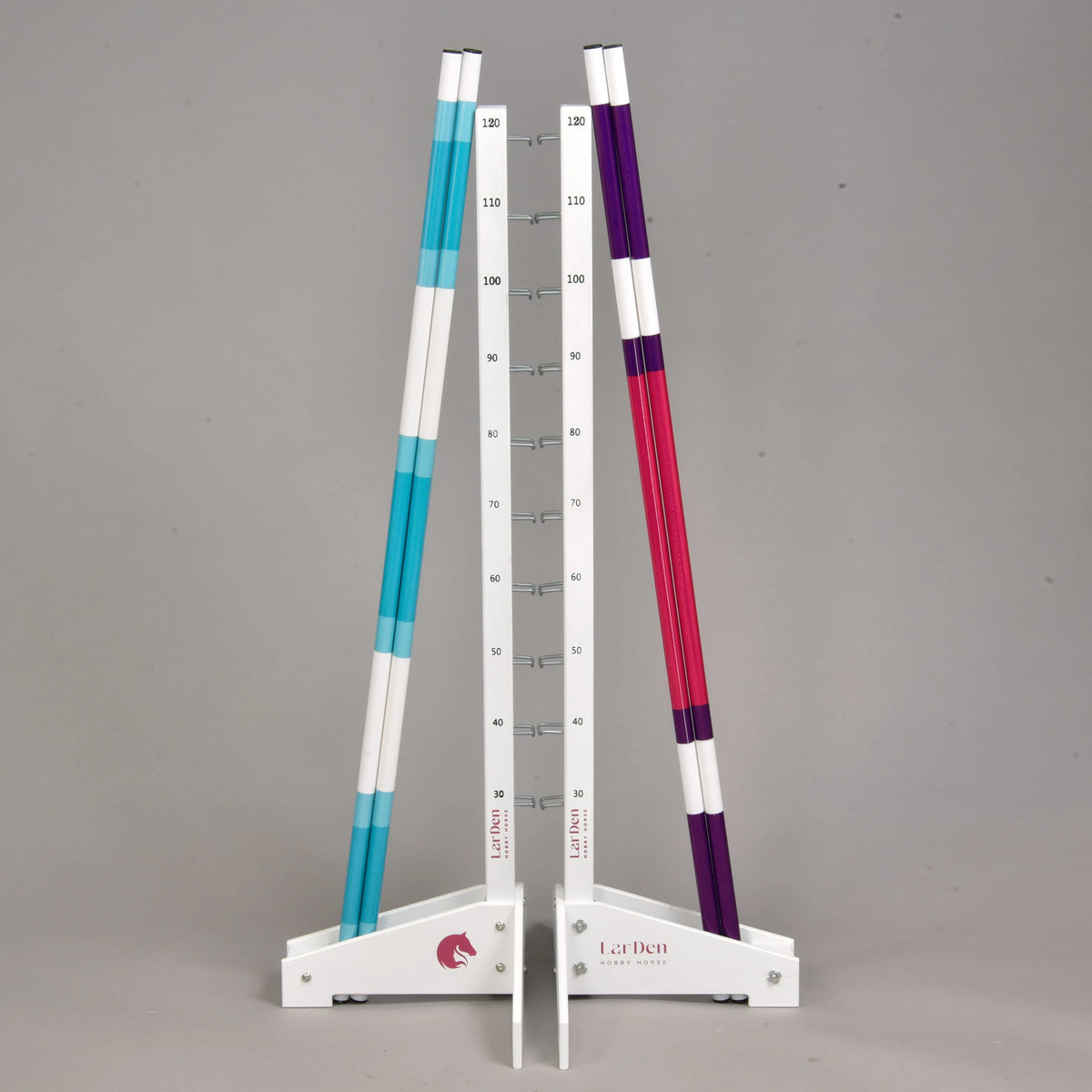 Set prekážky kolmák 120 cm + 2 ks bariérky od Hobby Horse LarDen s dvoma bielymi stojanmi so značkami merania, ktoré nesú tri pruhované tyče – dve modré a jednu fialovú. Táto nastaviteľná prekážka je vhodná na tréning Hobby Horsing vnútri aj vonku na sivom pozadí.