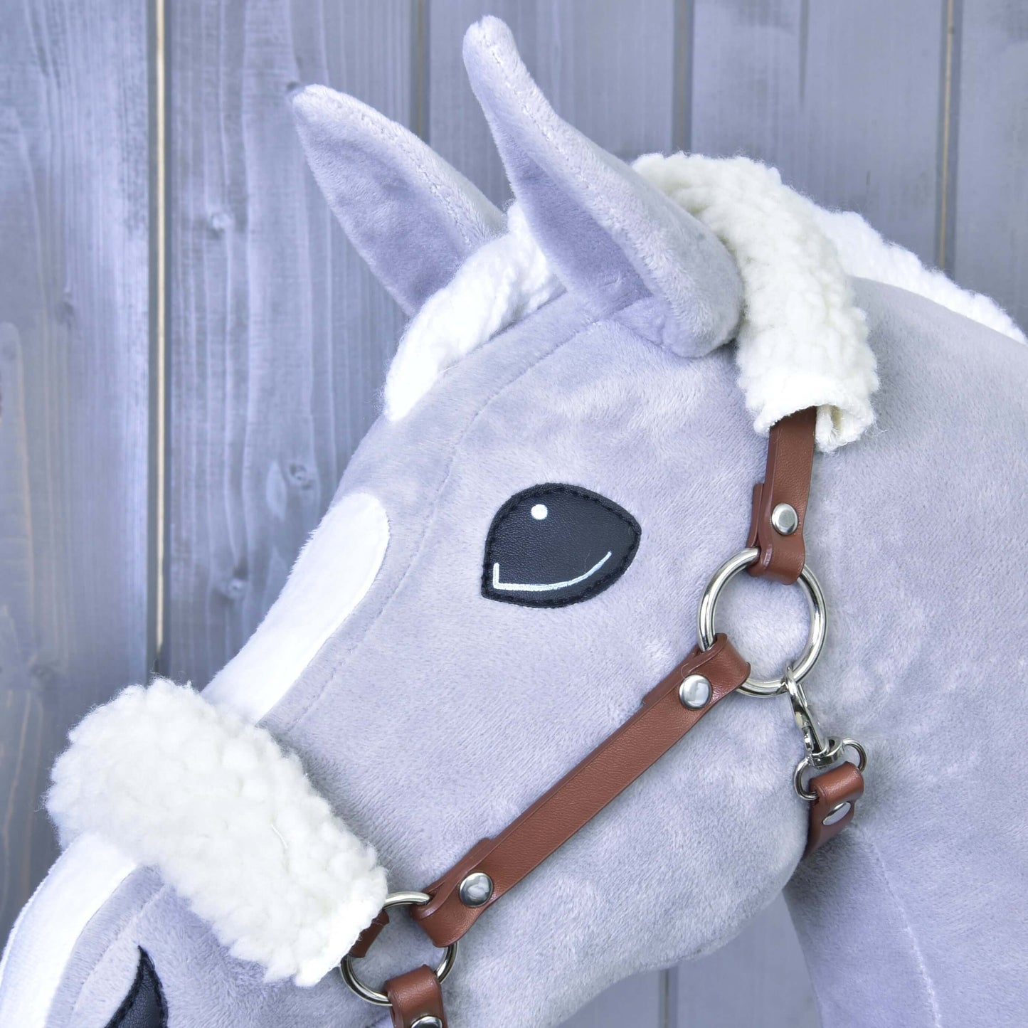 Detail plyšového koně Hobby Horse LarDen se světle šedým tělem, bílou hřívou a hnědou ohlávkou z koženky s beránkem na světlém dřevěném pozadí.