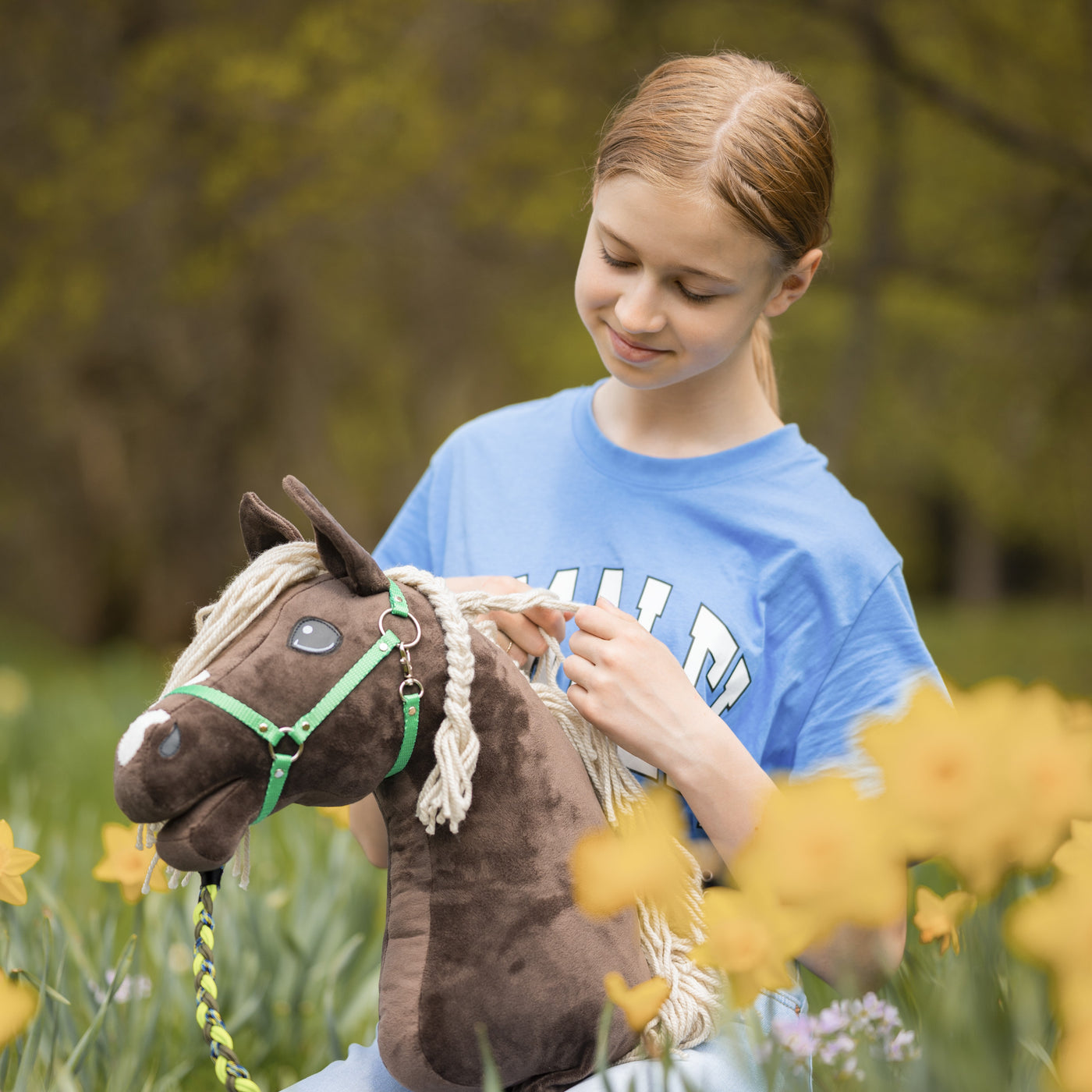 Dívka v modrém tričku se usmívá, zatímco drží a hladí svého hnědého Hobby Horse, sedí v poli žlutých narcisů a v pozadí jsou rozmazané zelené stromy.