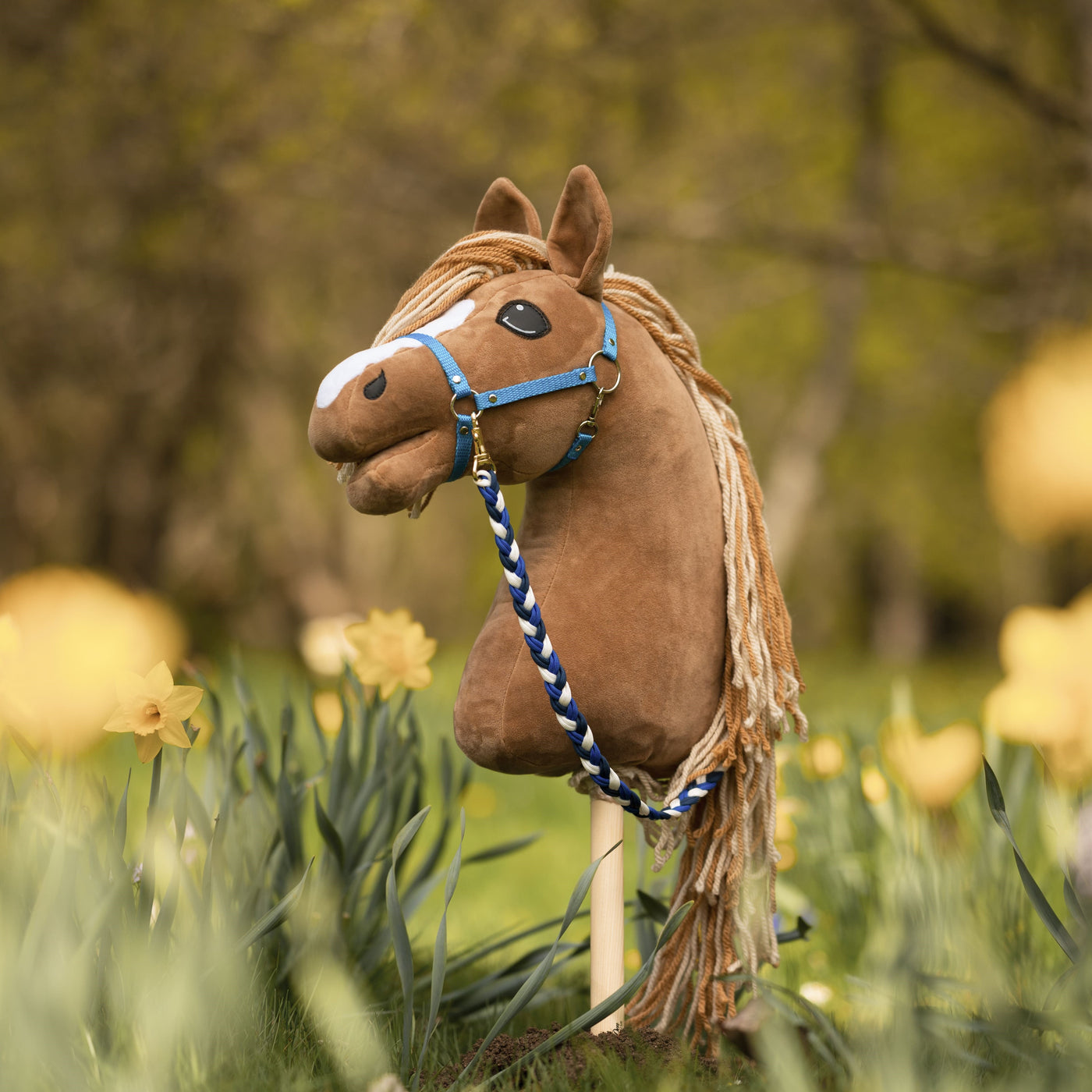 Hnědý Hobby Horse s modrou uzdečkou a přízovou hřívou stojí mezi žlutými narcisy v travnaté, sluncem zalité zahradě – dokonalá scéna pro Hobby Horsing s rozmazanými zelenými stromy v pozadí.