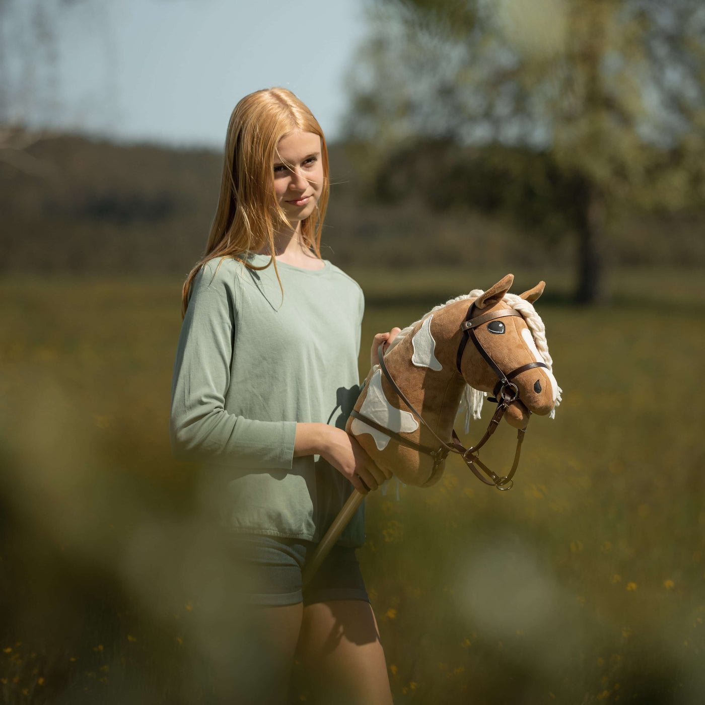Mladá dívka s dlouhými blond vlasy stojí venku na zelené louce a drží svého hnědobílého Hobby Horse – užívá si zábavu Hobby Horsing mezi stromy a rozostřenou zelení v pozadí.