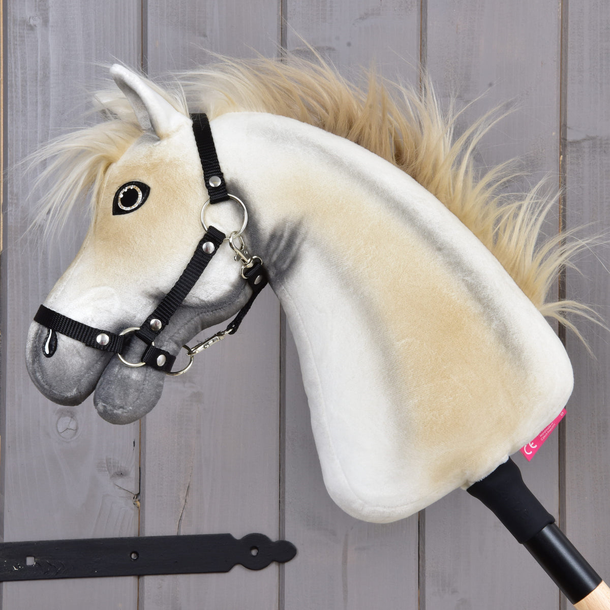 Hobby Horse Honey Pearl s čiernou ohlávkou - veľkosť S od Hobby Horse LarDen, béžovosivý plyšový koník na tyči s huňatou hrivou, čiernou ohlávkou a na tyči, vystavený pred drevenou stenou.