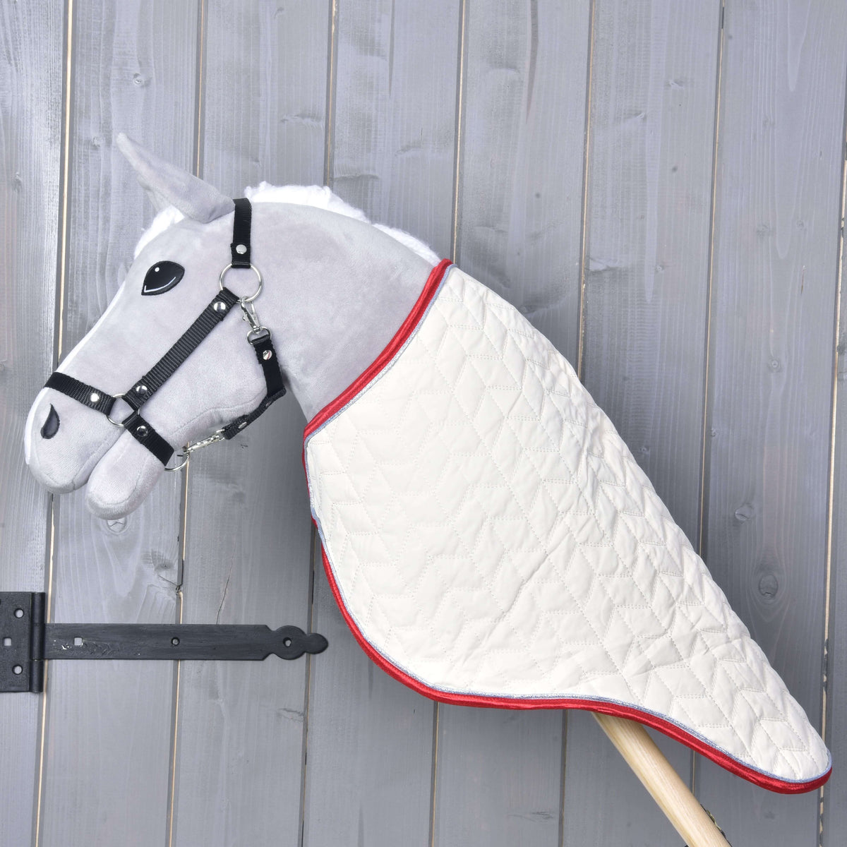Hobby Horse LarDen Prepravná deka Italy na sivom plyšovom Hobby Horse s čiernou uzdečkou a prešívanou bielou dekou, na drevenej tyči pred sivou drevenou stenou.