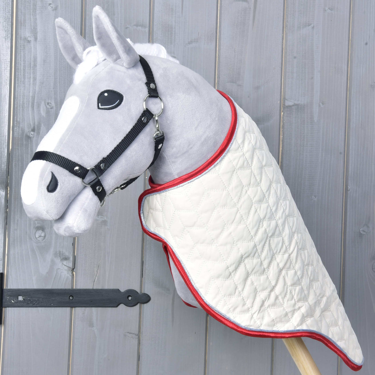 Prepravná deka Italy od Hobby Horse LarDen na sivom plyšovom Hobby Horse s čiernou uzdečkou; prešívaná biela deka s červeným lemovaním, na drevenej tyči pred svetlosivým dreveným pozadím.
