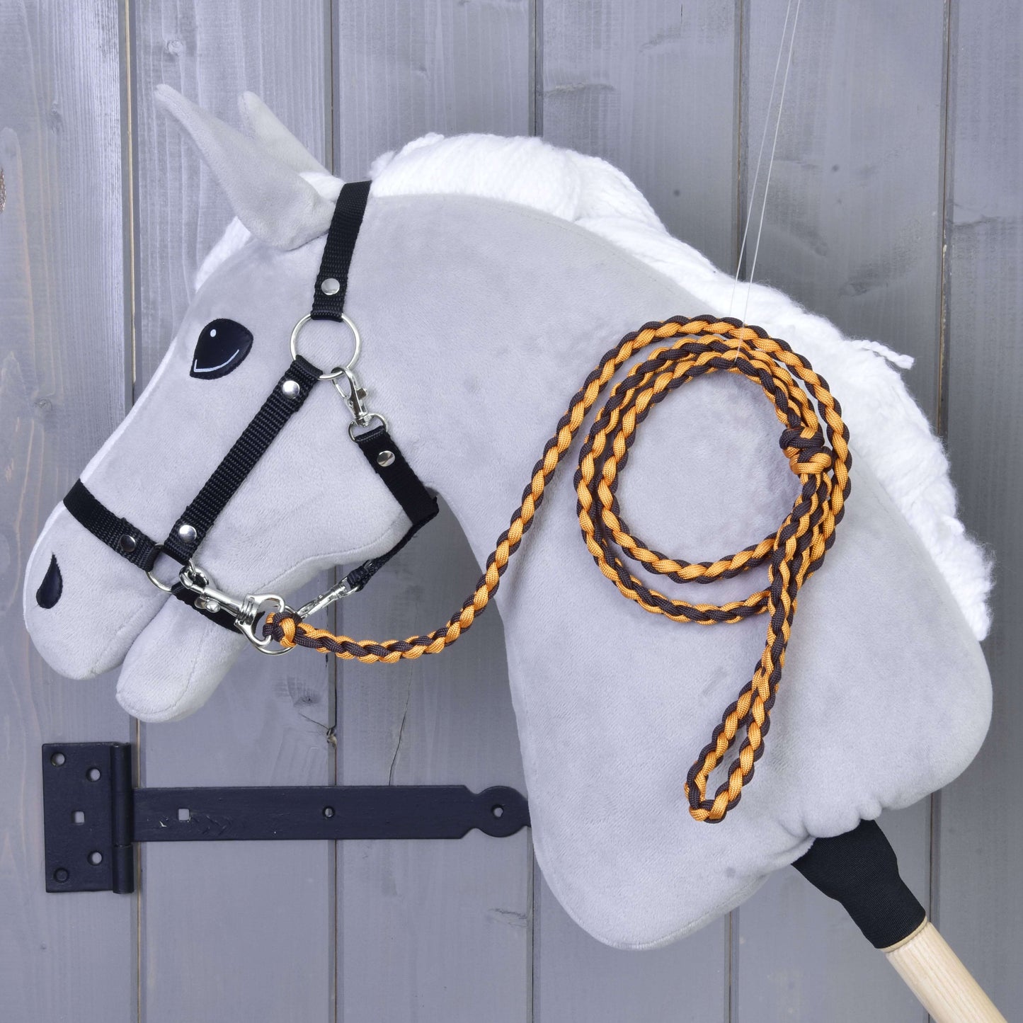 Hobby Horse - pletené vodidlo Pomello od Hobby Horse LarDen na plyšovom sivom Hobby Horse s bielou hrivou, čiernou uzdečkou a oranžovo-čiernym pleteným vodidlom s karabínou, pripevnenom na drevenej stene.