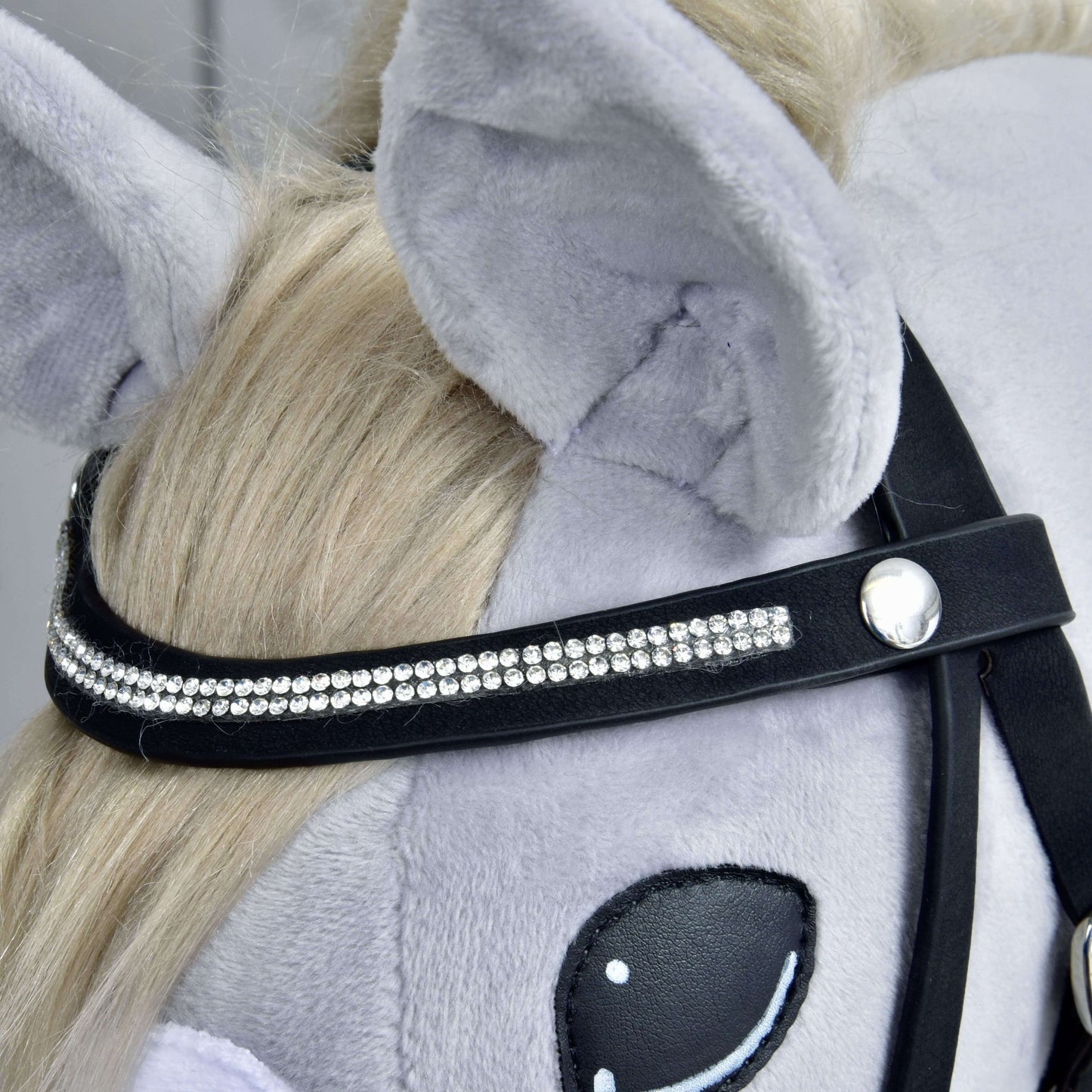 Detail hlavy plyšového koňa so svetlosivou srsťou, blond hrivou a kompletnou uzdečkou De Luxe s 2 čelenkami od Hobby Horse LarDen. Ozdobný pásik má lesklé kamienky a vidno detaily ucha a oka.