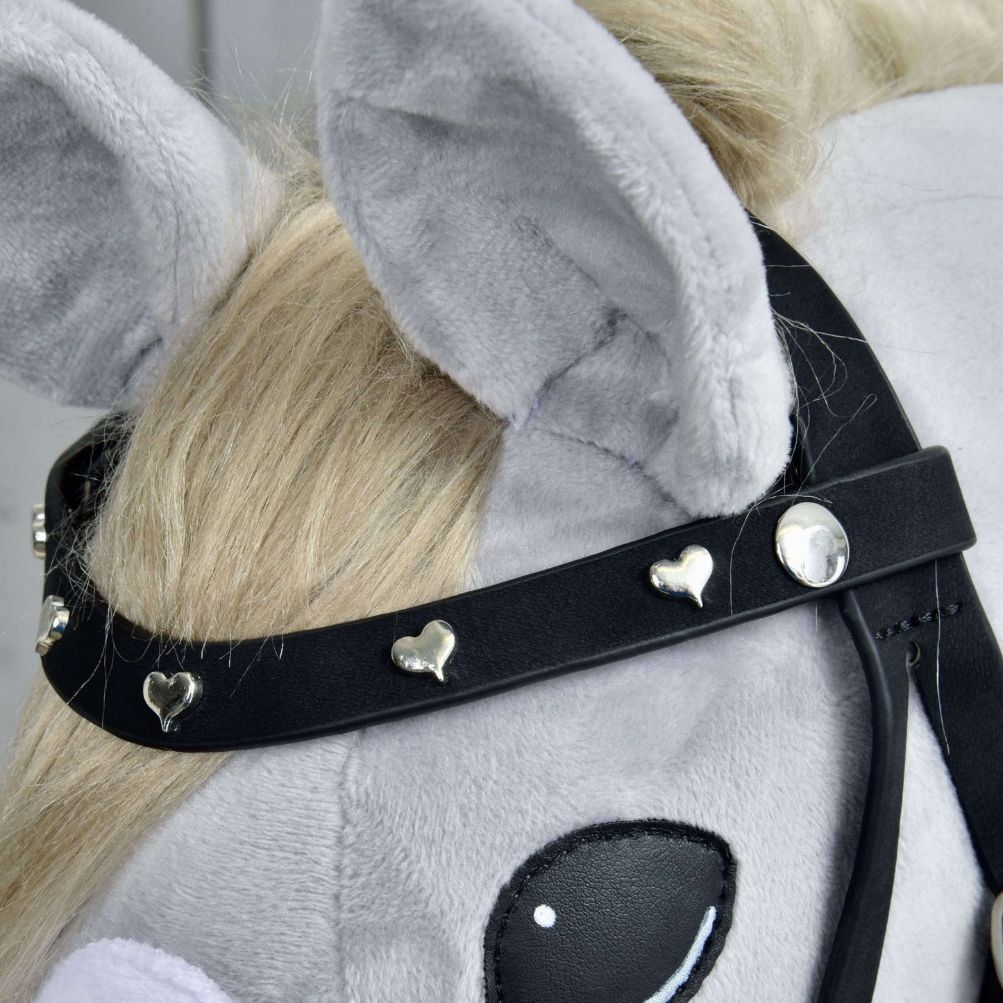 Detail hlavy sivého plyšového Hobby Horse s kompletnou uzdečkou De Luxe s 2 čelenkami od Hobby Horse LarDen, s dvoma odnímateľnými čelenkami zdobenými malými striebornými srdiečkami a okrúhlymi cvokmi. Kôň má svetlo blond hrivu a veľké mäkké uši.