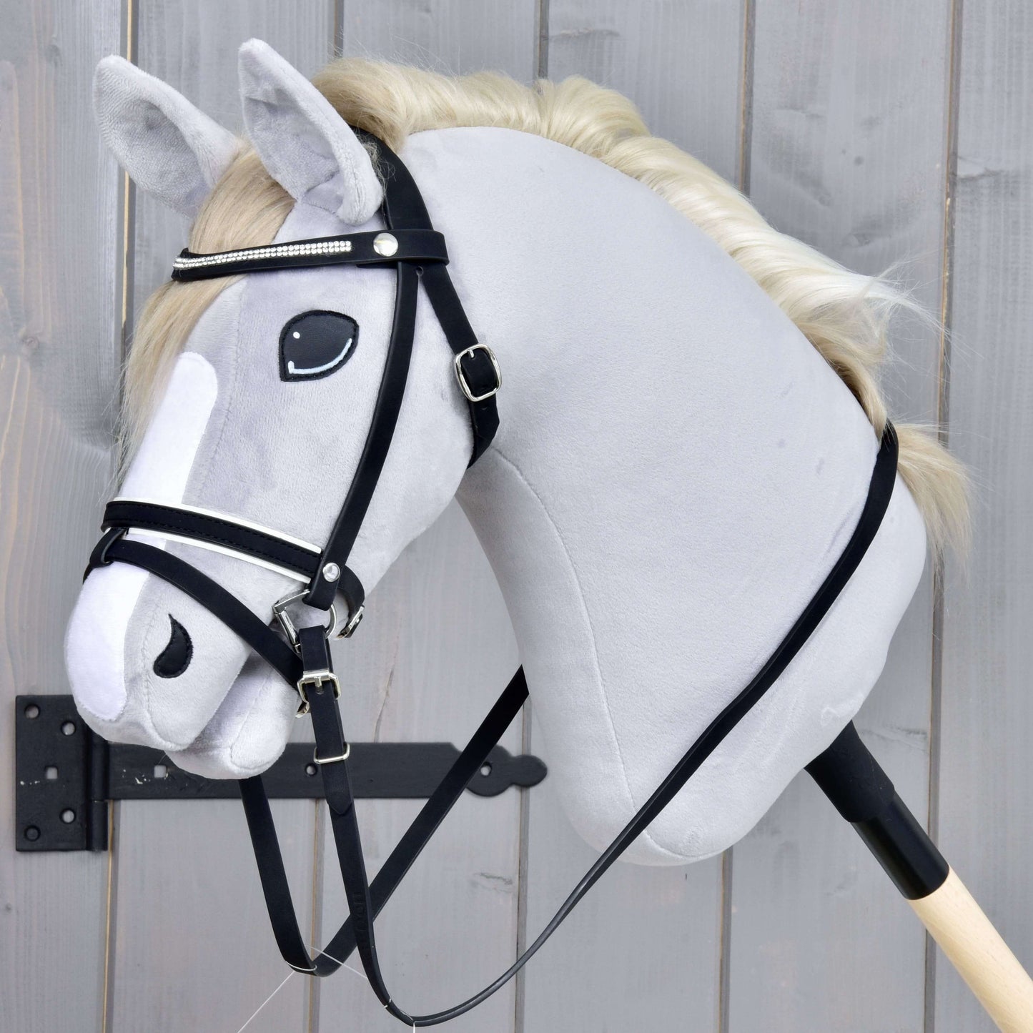 Kompletná uzdečka De Luxe s 2 čelenkami od Hobby Horse LarDen na realistickom sivom Hobby Horse na tyči s bielou hrivou a kombinovaným nánosníkom, nasadenom na drevenej tyči pred sivým dreveným pozadím.