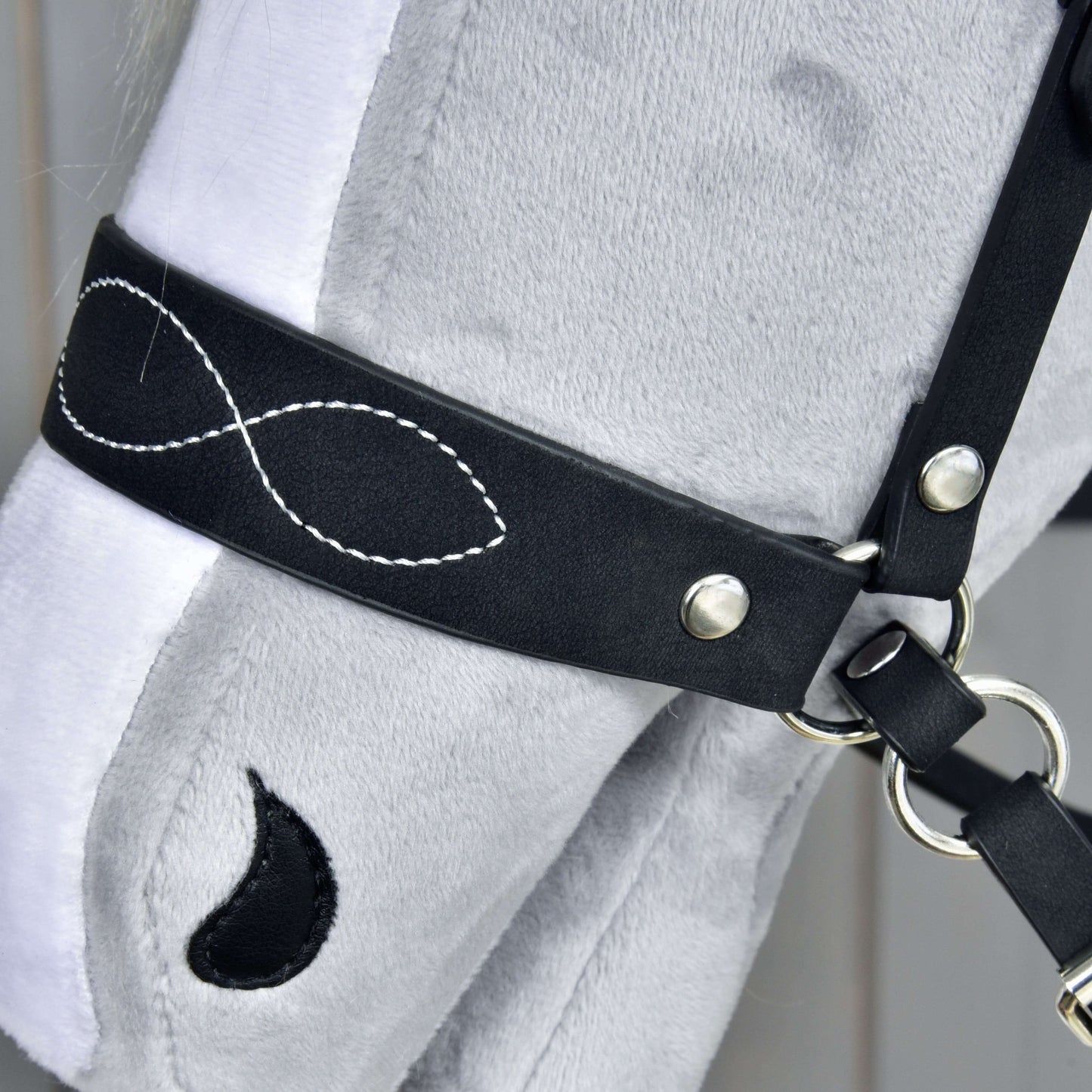 Detail hlavy sivého plyšového Hobby Horse s Kompletnou uzdečkou bezzubadlovou zdobenou od Hobby Horse LarDen, zdobenou bielym prešitím v tvare symbolu nekonečna a čiernou škvrnou v tvare kvapky pri oku.