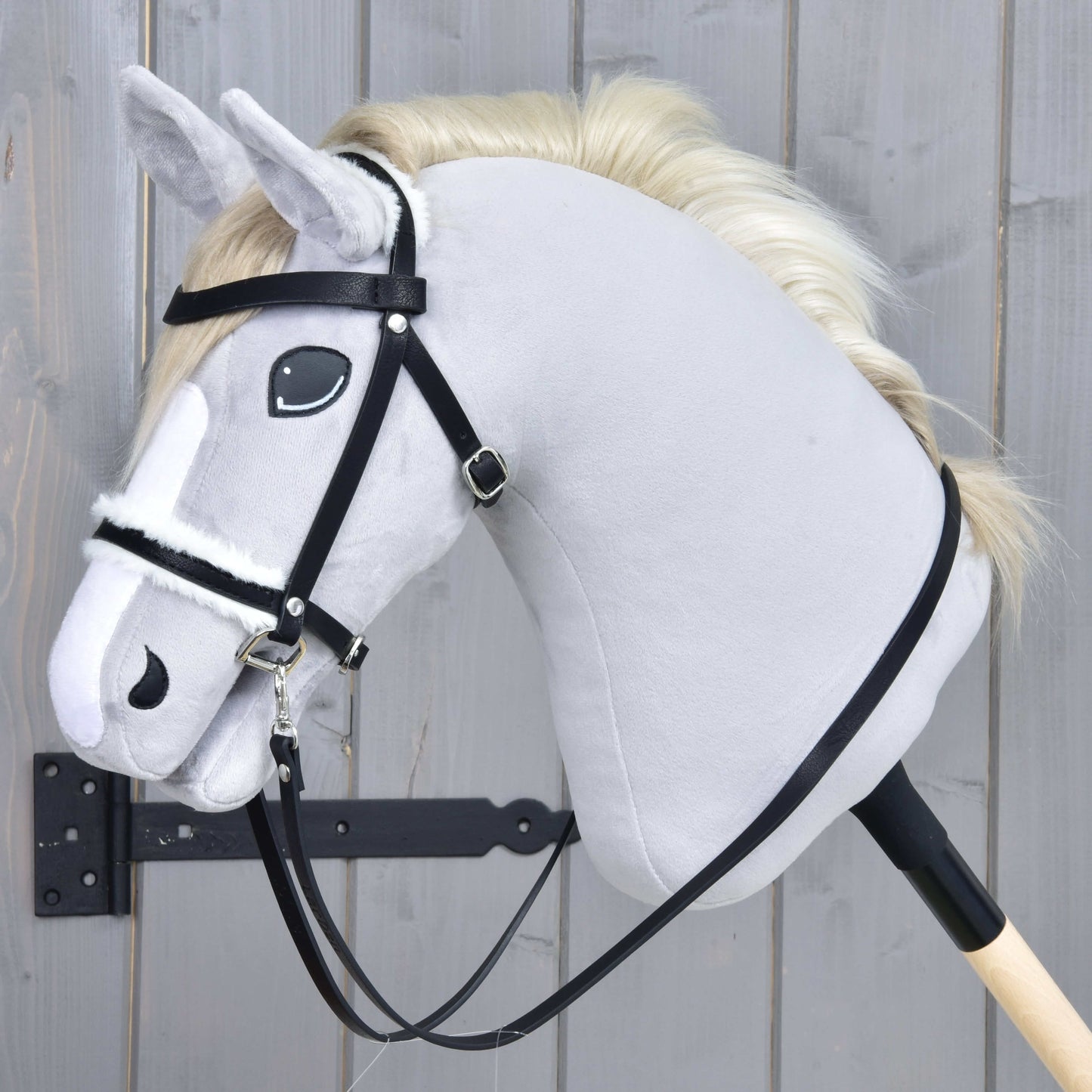 Kompletná uzdečka s barančekom čierna od Hobby Horse LarDen na svetlosivom Hobby Horse s krémovou hrivou, s čiernou koženkovou uzdečkou a opratami na drevenej tyči, na pozadí sivého dreva.