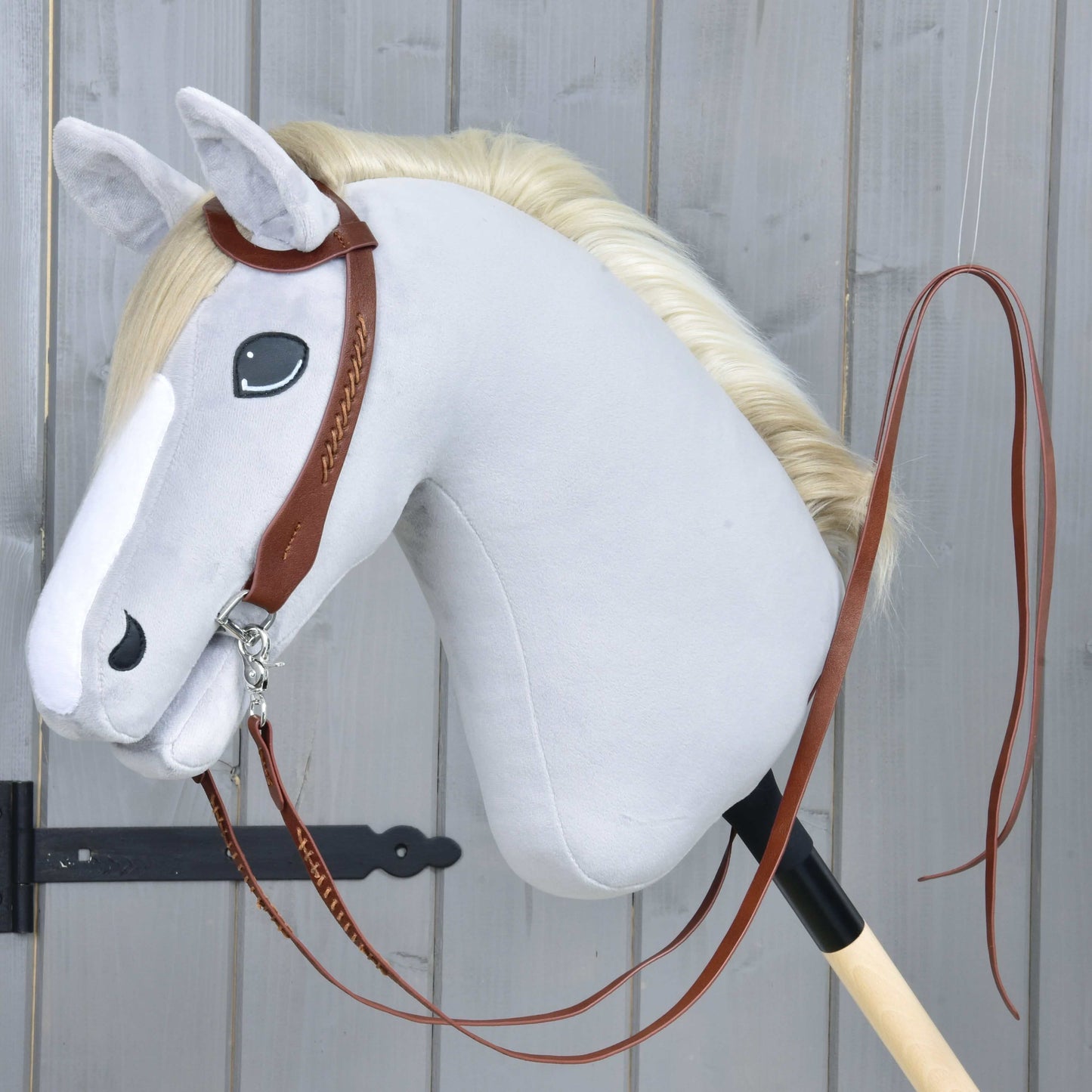 Plyšový Hobby Horse od Hobby Horse LarDen so svetlosivou hlavou, bielou hrivou a Kompletnou westernovou uzdečkou jednouška z kože s delenými opratami, upevnený na drevenej tyči pred sivou drevenou stenou.