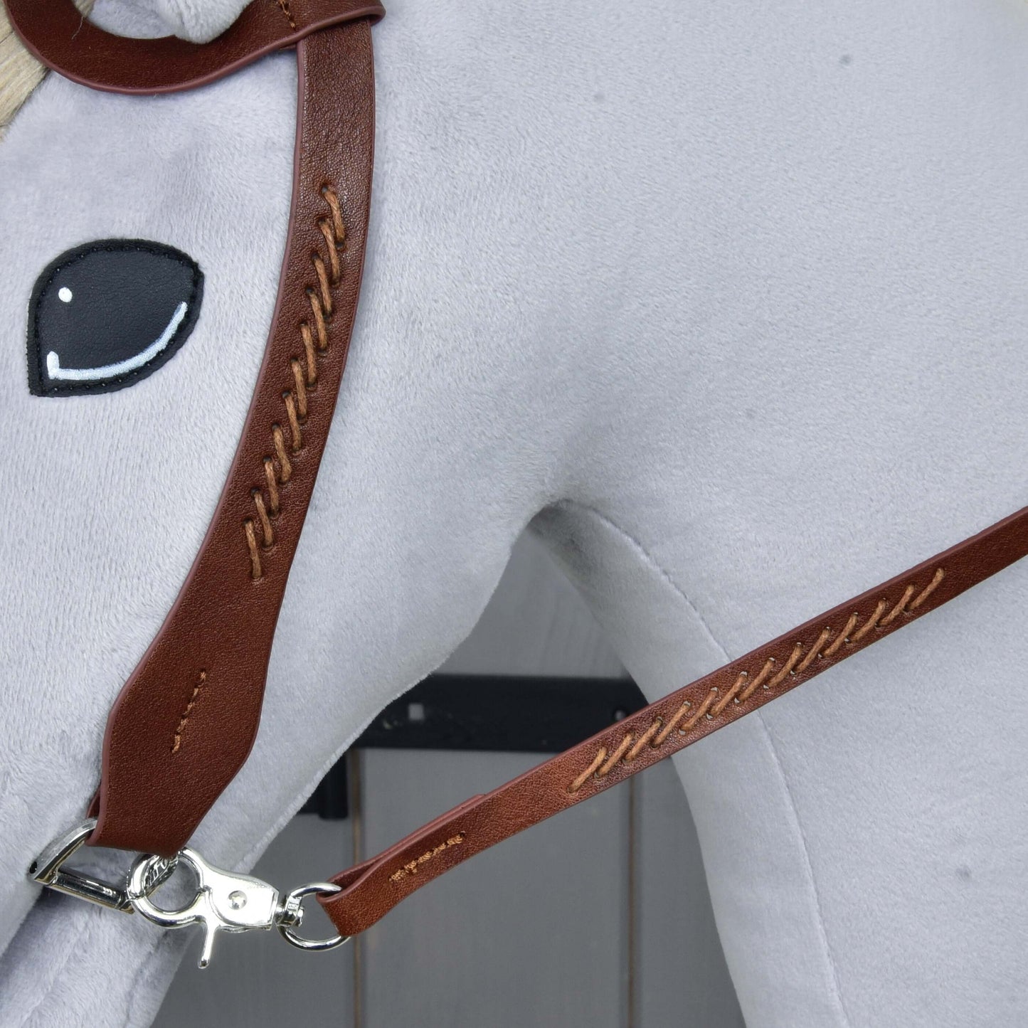 Detail plyšovej konskej hlavy Hobby Horse LarDen s Kompletnou westernovou uzdečkou jednouška z kože z hnedej kože, s výrazným prešívaním, delenými opratami, kovovou sponou a jednoduchým čiernobielo vyšitým okom na svetlosivom pozadí.