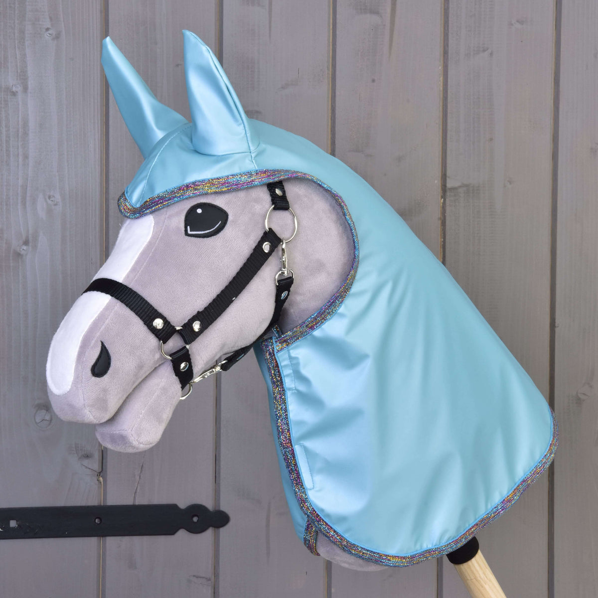 Sivý Hobby Horse od Hobby Horse LarDen s čiernou uzdečkou, veľkými plstenými očami a kapucňou Pláštenka Light Blue s ladiacimi ušami, zobrazený pred drevenou stenou.